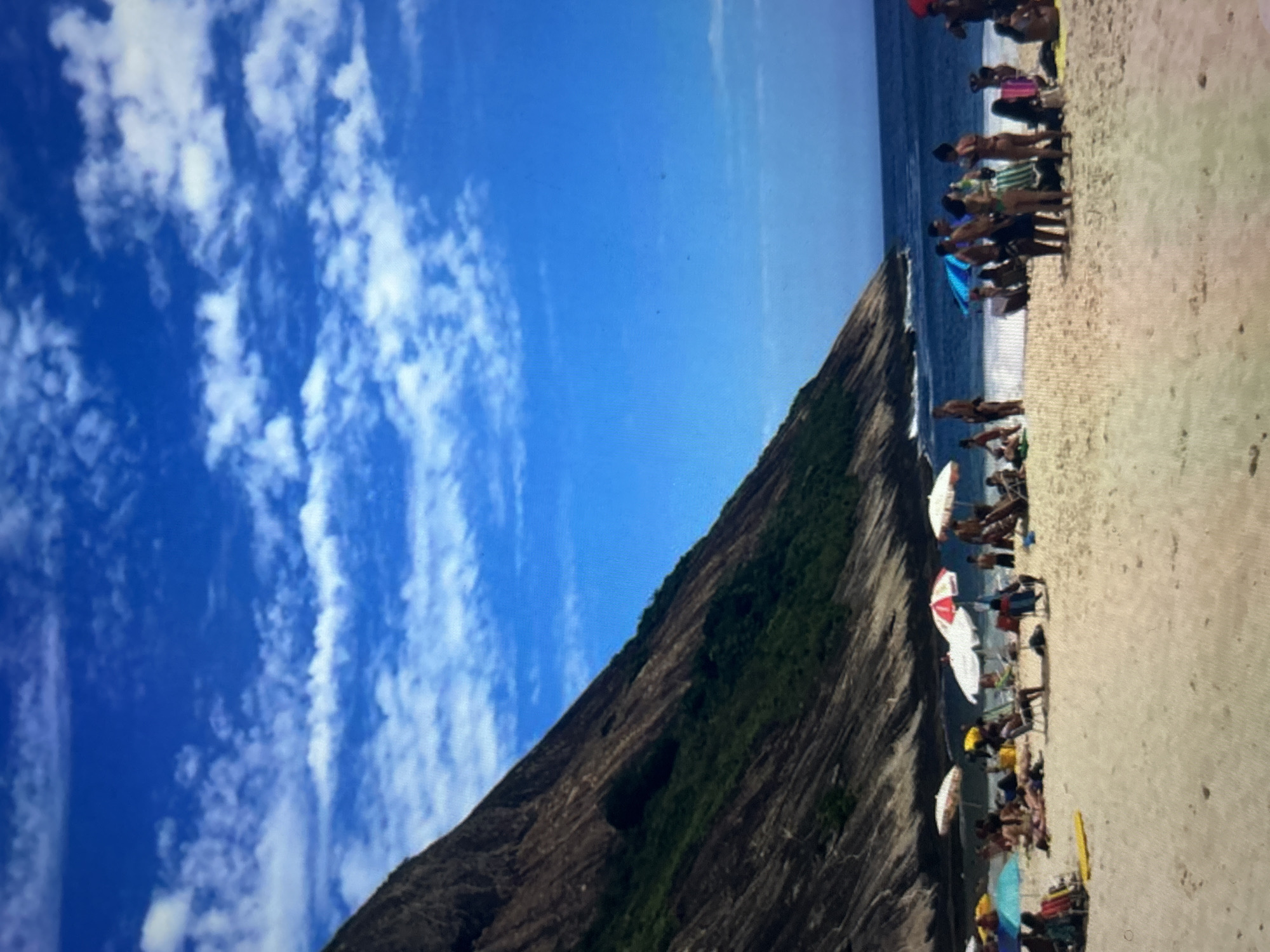 The image depicts a sunny day at a crowded beach. A steep, rocky mountain covered in green vegetation dominates the left side of the frame, meeting a sandy beach where numerous people are gathered. The ocean stretches out beyond the beach, with gentle waves lapping at the shore.
The beach is filled with individuals sunbathing, socializing, and relaxing under colorful umbrellas. Beach chairs and towels are scattered across the sand. The sky is a vibrant blue, dotted with fluffy white clouds, suggesting fair weather. The lighting indicates it is likely daytime, possibly mid-morning or afternoon, given the bright sun.
The scene appears to be a popular tourist destination, with people of various ages enjoying a day outdoors. The atmosphere seems relaxed and leisurely, typical of a beach outing.
Notable details include the contrast between the rugged mountain and the serene beach, the array of beach umbrellas providing shade, and the clear blue sky. There is no visible text in the image.