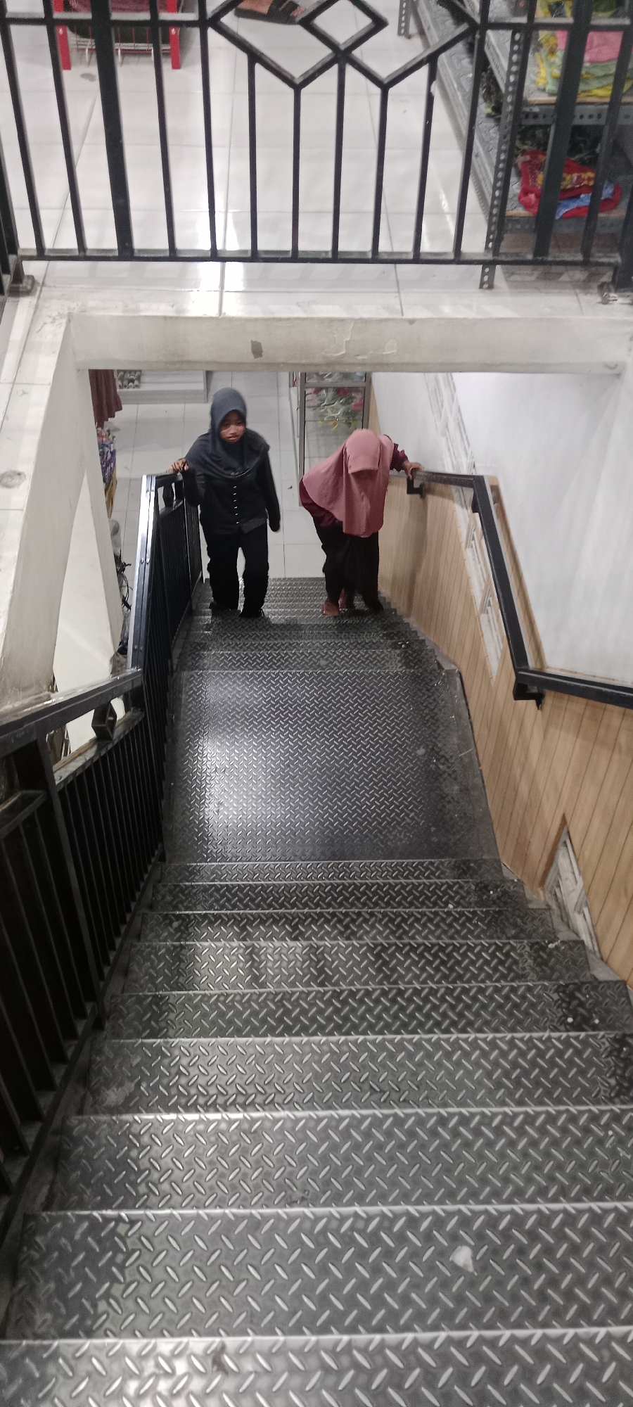Two young females, wearing hijabs and modest clothing, are descending a metal staircase in a building. The female on the left is wearing a dark grey hijab and a black jacket over dark pants, and she is holding onto the black metal railing. The female on the right is wearing a reddish-brown hijab and a dark red top with dark pants, and she is also holding onto the railing. The staircase has a diamond plate pattern on the treads. The walls of the stairwell are lined with light brown wood paneling, and the steps are illuminated by overhead lighting which casts shadows. The location is identified as Rengel, Indonesia.