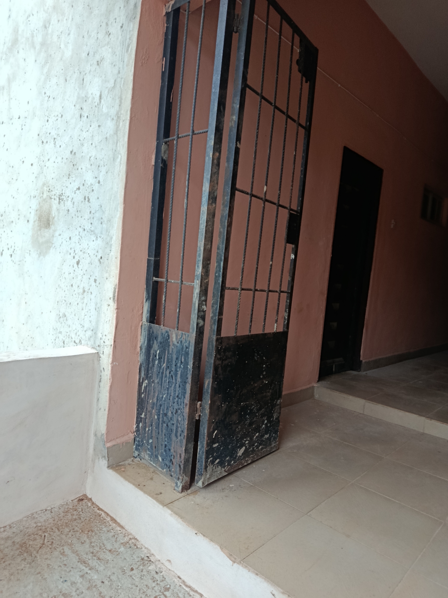 The image depicts an exterior view of a building in Ukpor, Nigeria, with a focus on a partially open, black metal security gate. The gate is positioned in the foreground, leaning inward and appearing somewhat weathered and dirty. To the left of the gate, a textured, off-white wall shows signs of discoloration and speckled markings. The wall to the right of the gate is painted a muted reddish-orange and extends down a hallway. Further down the hallway, a dark doorway or opening is visible, suggesting an entrance or passage. The floor in front of the gate is tiled in a light beige color, with some areas appearing worn or dusty. A raised white ledge runs along the bottom of the off-white wall. The scene appears to be an outdoor corridor or entryway.