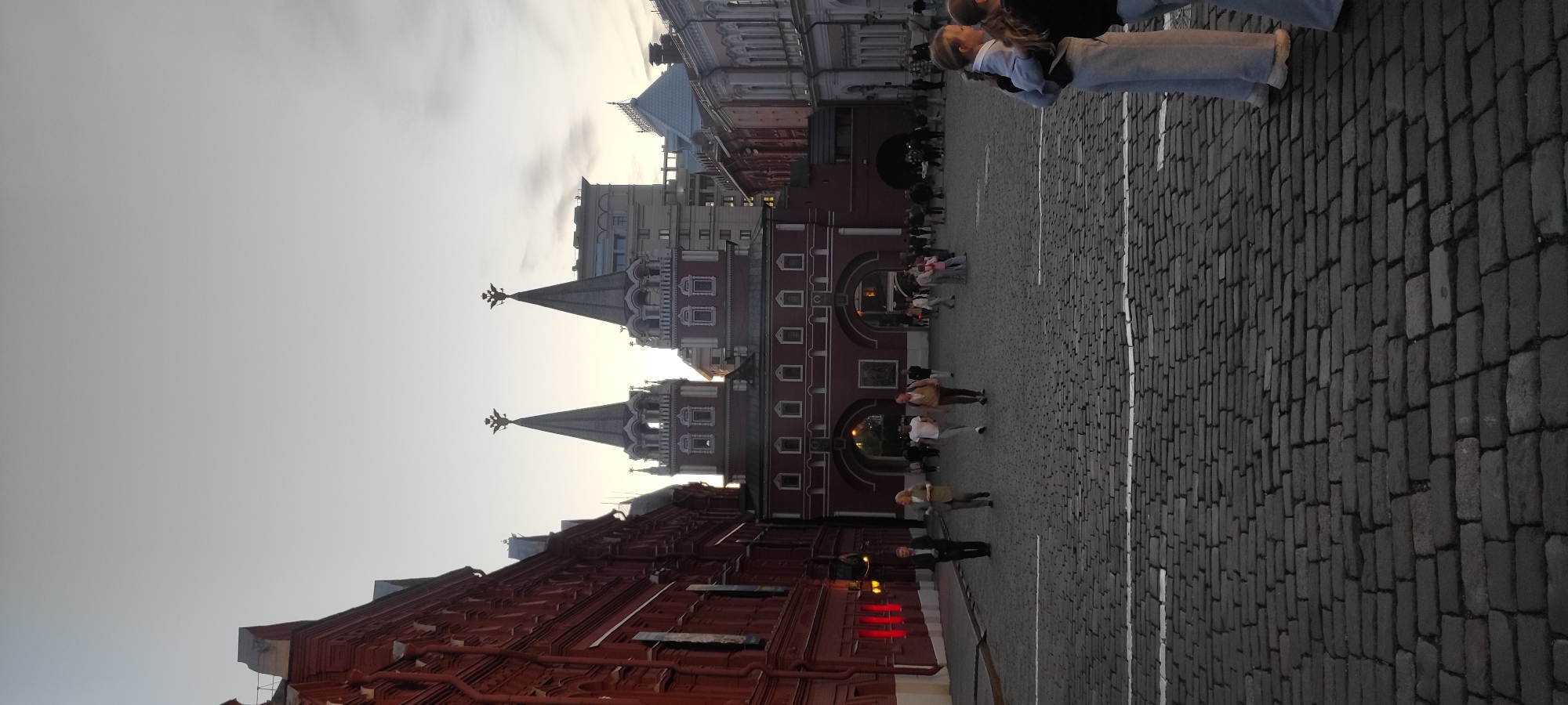 The image captures an outdoor scene on a cobblestone plaza in front of a red brick building with arched entrances and two prominent spired towers, suggesting a historical or governmental location. The sky is overcast, indicating a diffused light source, possibly early morning or late afternoon. Several people are scattered across the plaza, some walking individually or in small groups, while others appear to be standing and observing. A red brick building with multiple windows occupies the left side of the frame, and other architectural structures with a lighter colored facade are visible in the background. The ground is paved with dark, uneven cobblestones. The overall atmosphere appears to be calm and public, with people engaged in casual movement.
