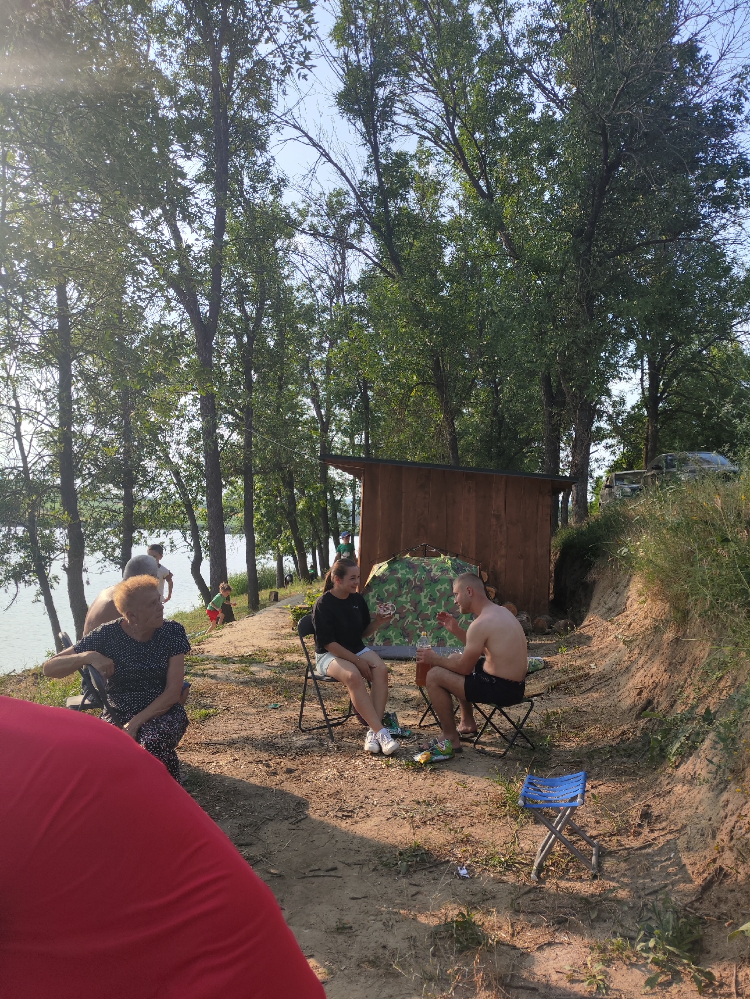 The image depicts a group of people at a waterfront location, likely a lake or river, in Găleștii Noi, Moldova. The scene is set on a sandy bank lined with trees. In the foreground, a person wearing a red garment is partially visible. To the left, an older woman with curly orange hair is seated in a black folding chair, wearing a black and white polka dot dress and patterned pants. She is looking towards the left. Beside her, partially obscured, is another person. Further back, a man and a woman are seated on folding chairs. The woman, wearing a black top and denim shorts, is looking to her left and smiling. The man, shirtless and wearing black shorts, is looking at the woman and holding a bottle. A camouflage tent is pitched behind them, and a small wooden structure resembling a shed is situated to the right of the tent. In the background, more trees line the water, and a few vehicles are visible further up the bank. A child in a red shirt and green shorts is seen playing near the water. The overall impression is a casual outdoor gathering or picnic.