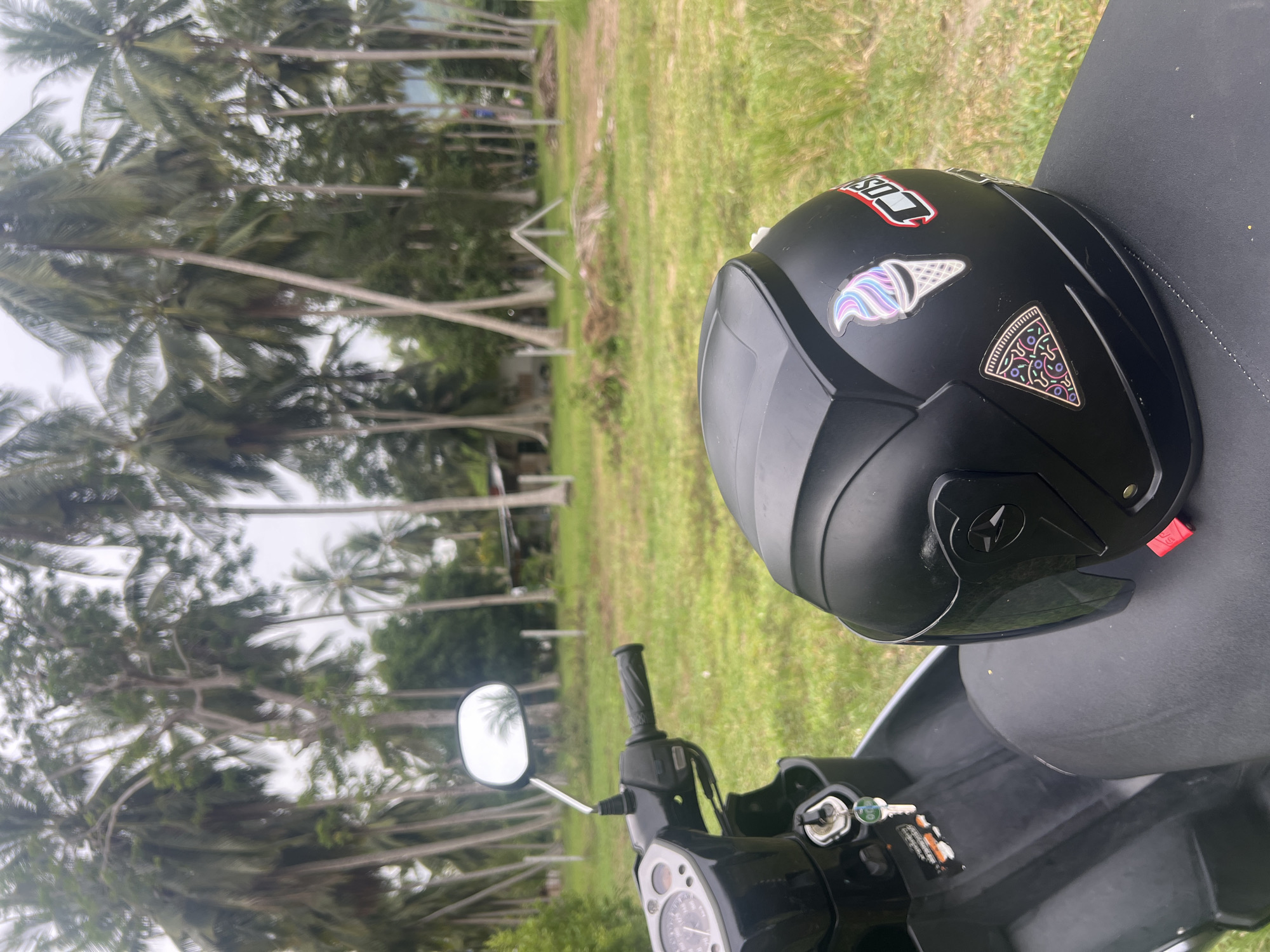 A close-up shot shows a black motorcycle helmet resting on the seat of a black motorcycle. The helmet is adorned with stickers: one depicts an ice cream cone in pastel colors, and another shows a slice of pizza. To the left of the helmet, the motorcycle's handlebars and instrument panel are visible, with keys in the ignition.
The motorcycle is parked on a grassy area, likely a rural roadside. In the background, a dense grove of tall palm trees creates a tropical atmosphere. Beyond the trees, a modest, weathered building can be seen, along with some fencing and what appears to be agricultural land. The sky is overcast, suggesting a diffused light. The overall scene evokes a sense of casual travel in a warm, possibly remote, tropical location. The location is specified as Tambon Ko Phangan, Thailand.