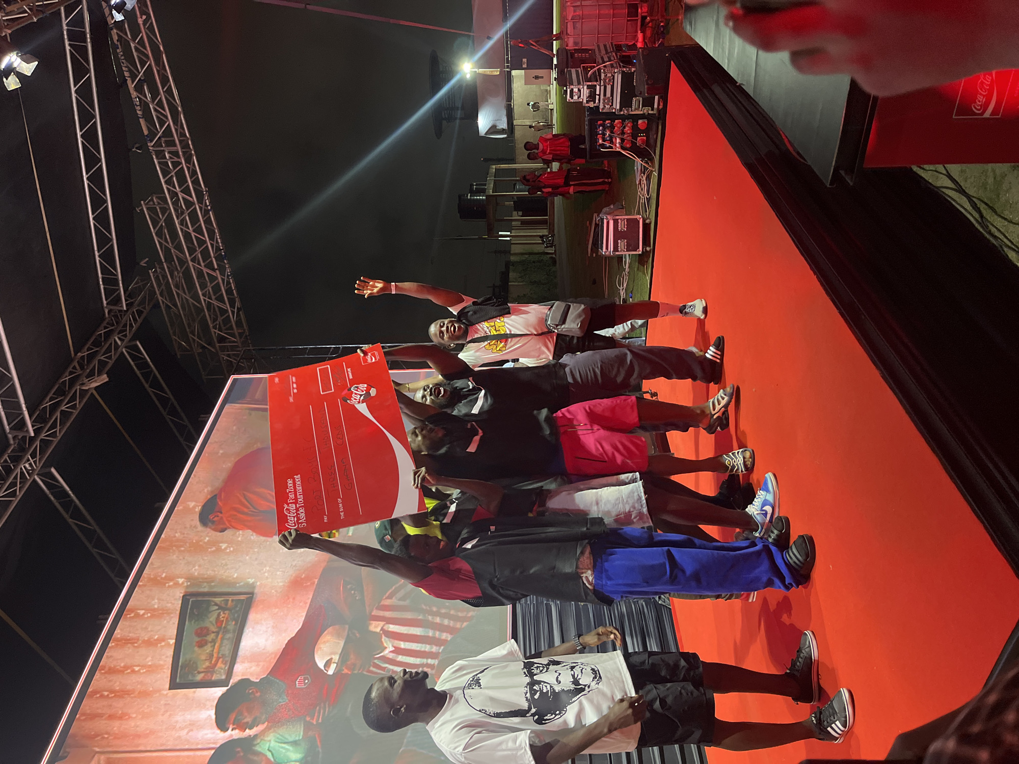 In Accra, Ghana, a group of young men on a stage celebrate winning a Coca-Cola Fan Zone 5-A-Side Tournament. They are holding up a large red novelty check with "Coca-Cola Fan Zone 5 Aside Tournament" printed on it. The check has handwritten text that appears to be "PORT BOY..." for the payee and "GHANA CANS" for the sum. The men are dressed casually in t-shirts, shorts, and sneakers. Some are raising their hands in the air, while others are smiling and looking at the camera. The stage is covered in red carpet, and behind them, a large screen displays an image of people in red and white jerseys. The event appears to be taking place outdoors at night, with stage lighting and scaffolding visible in the background. In the far background, there are buildings and some spectators.