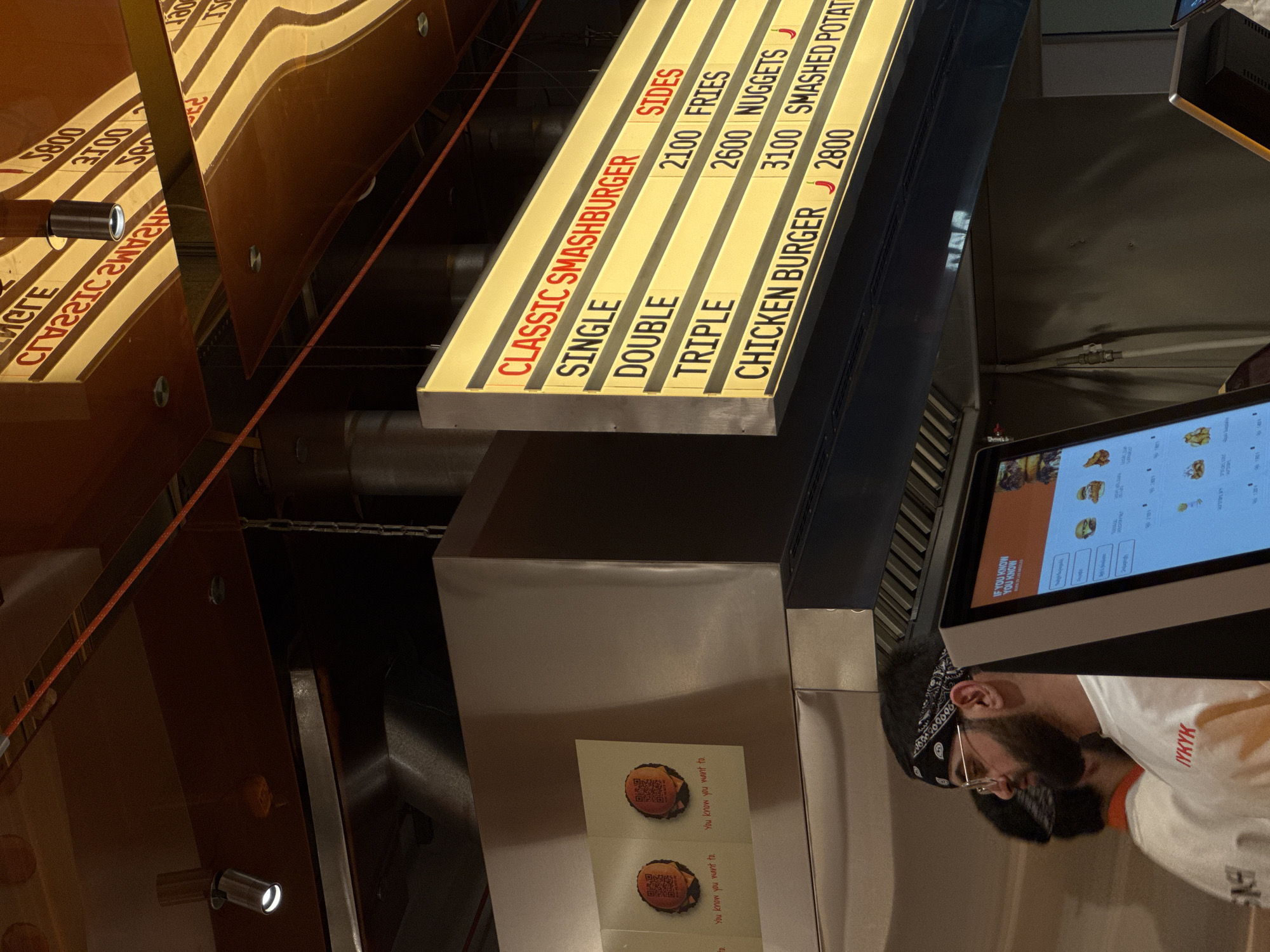 The image shows a portion of a restaurant interior, likely a fast-food establishment. The menu board advertises "CLASSIC SMASHBURGER" options, including single, double, and triple burgers, as well as sides like fries, nuggets, and smashed potatoes. A "CHICKEN BURGER" is also listed. A person is seen partially in the frame, wearing glasses and a bandana. A digital menu is visible in the foreground. The environment appears to be a service area where orders are placed. It is difficult to know the specific location in Yerevan, Armenia without more context.