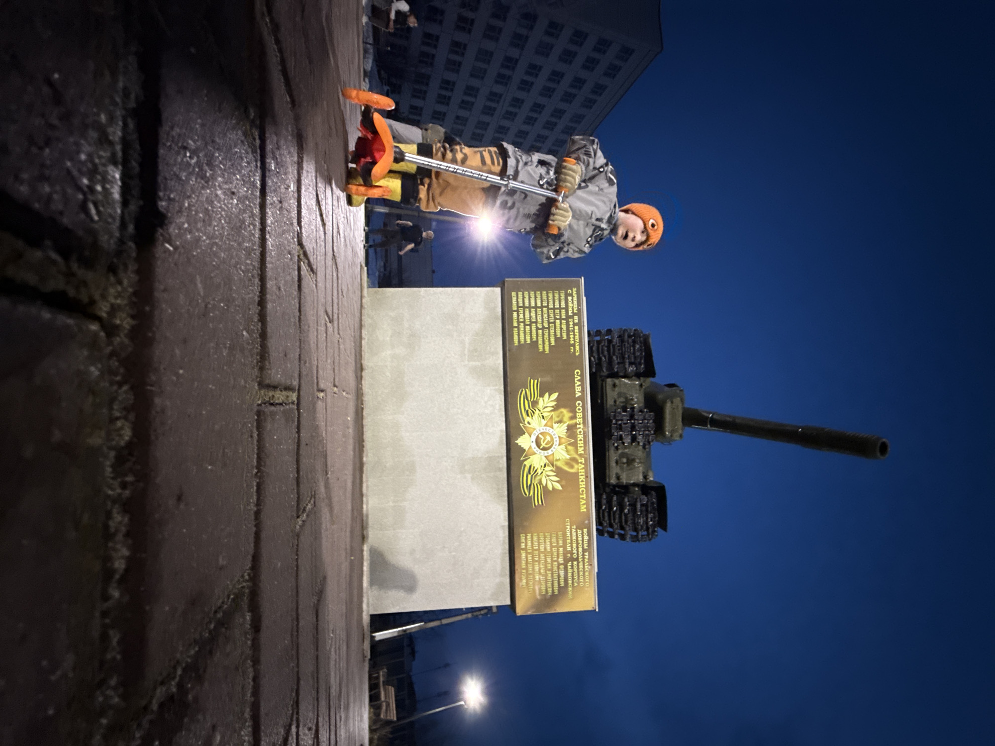 The photo shows a young boy riding a scooter on a brick-paved area next to a tank monument at night. The monument consists of a concrete pedestal supporting a T-34 tank. A plaque on the pedestal commemorates Soviet tank crews, specifically those from the Ural Volunteer Tank Corps, who did not return from the 1941-1945 war. The plaque also lists names of those who perished. The boy is wearing a winter hat, gloves, and a jacket. In the background, a large building is visible, along with street lights. The location appears to be the city of Chaikovsky, Russia.