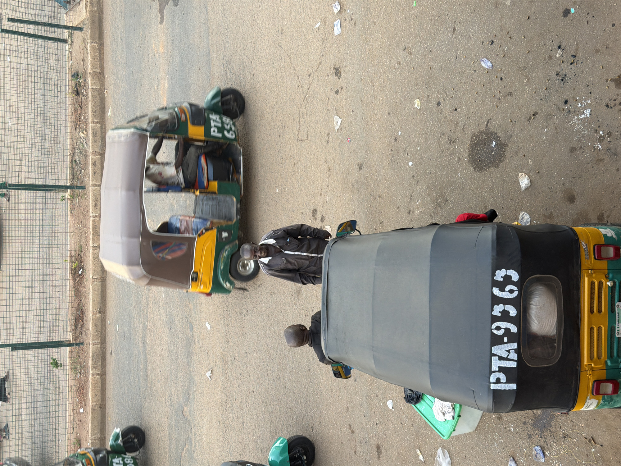 This is an overhead, landscape-oriented photograph taken in Bukuru, Nigeria. The scene depicts two auto-rickshaws, commonly known as Keke Napeps, parked on a paved road. The auto-rickshaws are primarily yellow and green, with gray canopies. One auto-rickshaw in the foreground has "PTA-9363" written in white on its side. The other auto-rickshaw, further back and to the left, has "PTA 659" written in white on its side.
Two men are visible. One man, wearing a dark jacket, stands next to the auto-rickshaw in the foreground. Another man, wearing a dark jacket, is seated in the driver's position of the auto-rickshaw in the background. The road surface is asphalt, with scattered debris and litter. To the left of the auto-rickshaws, a chain-link fence runs along a grassy embankment. The lighting suggests it is daytime, possibly late morning or early afternoon, with overcast conditions given the lack of harsh shadows. The overall impression is a common street scene in a Nigerian town.