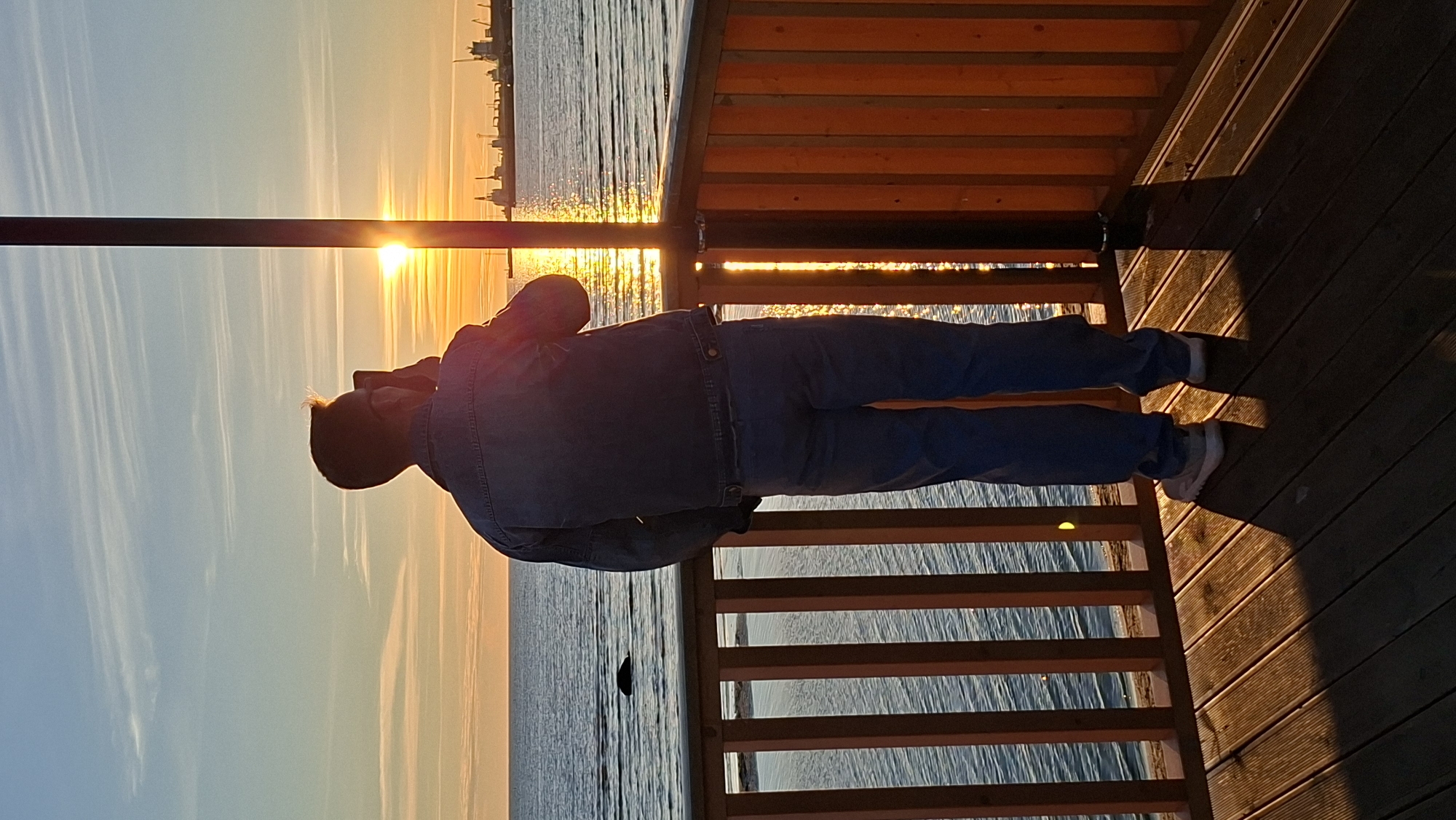 A person, viewed from the back, stands on a wooden pier, holding a phone to their ear, looking towards the sea. The sun is setting over the water, casting a warm glow across the scene. Ships are visible in the distance, on the horizon. The pier has wooden railings. The water is calm with gentle waves. The sky is a gradient of blue and orange, with wispy clouds. This scene is consistent with locations along the Tallinn coastline, possibly near the harbor area.