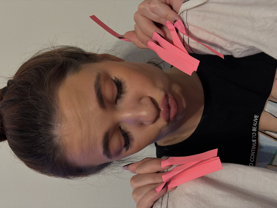 A woman is shown from the head up, lying down with her eyes closed. She has long eyelashes and her hair is pulled back. The woman's nails are long and pink paper strips are affixed to them, creating an extended artificial nail effect. She wears a black shirt with white writing that says "CONTINUE TO BE ALIVE". A white fabric is visible around her shirt. The picture appears to be taken with an overhead camera angle.