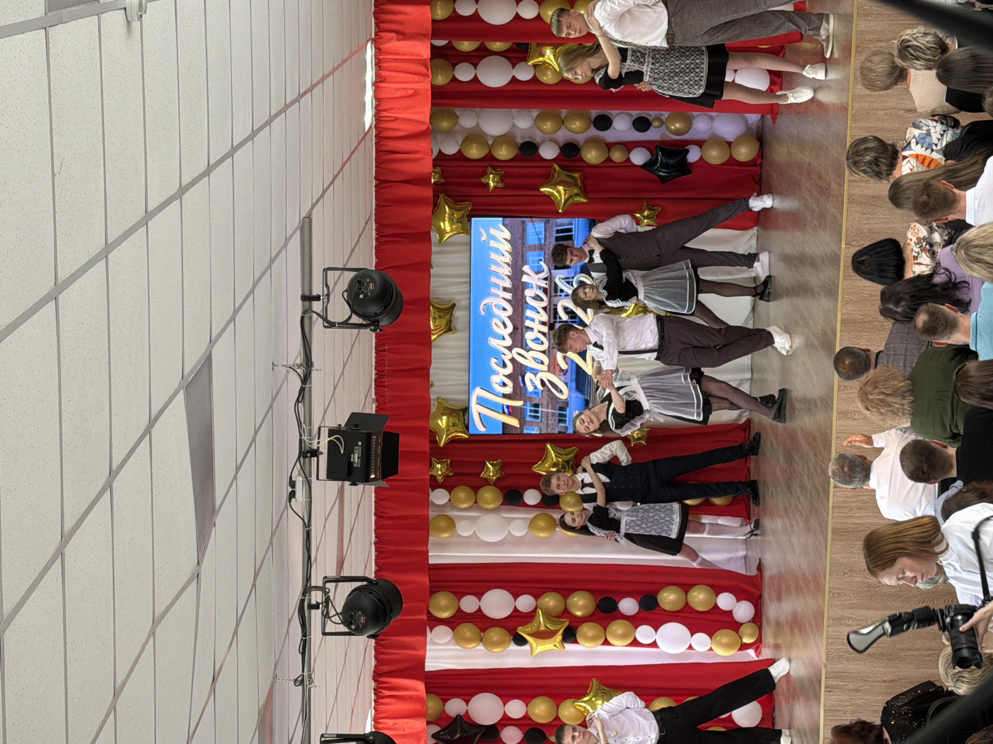 The image shows a stage setting in Izhevsk, Russia, likely for a school event. The stage is decorated with red and white fabric, gold and white balloons, and star-shaped decorations. A screen displays the Russian text "Последний звонок 2023," which translates to "Last Bell 2023." Several high school students are on the stage, some wearing traditional outfits. An audience is visible on the right side of the image, and lighting equipment is positioned on the left. A person with a camera is taking photos of the performance.
