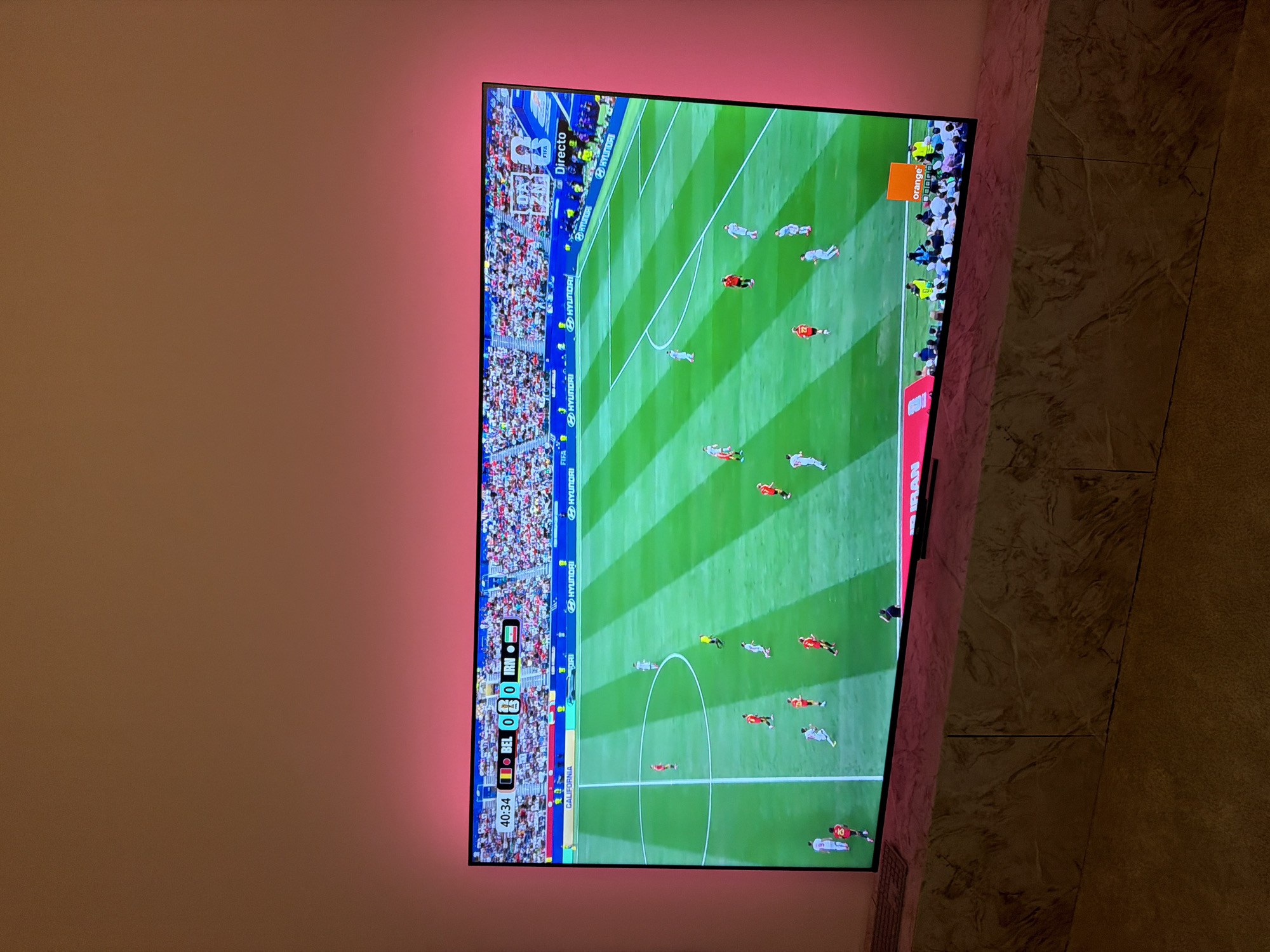 The image shows a television screen displaying a soccer match, specifically Belgium (BEL) versus Iran (IRN), with the score at 0-0 in the 40th minute. The TV is mounted on a wall in a domestic setting, with ambient pink lighting behind the screen. The room features a marble-patterned surface below the television, possibly a TV stand or shelf. The soccer field on the screen is vibrant green with white markings, and players in red and white uniforms are visible. Advertisements for Hyundai and California are displayed along the perimeter of the field. The overall scene depicts a modern living space with entertainment technology. There is no indication that this location is in Barcelona, Spain; it appears to be a generic indoor environment.
