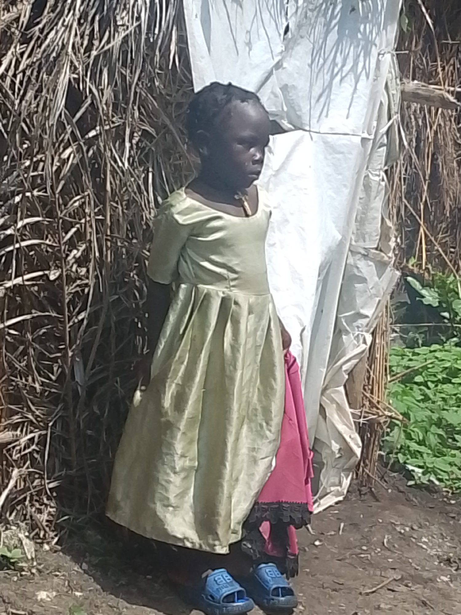 A young child stands outdoors near a structure made of dried plant material and white sheeting, with green vegetation in the background. The child is wearing a light green dress with pleats and a pink garment peeking out from underneath, along with blue sandals. The child's head is turned to the right, and they appear to be looking downwards. The ground is a mixture of dirt and scattered debris. The scene is set in Otulu, Nigeria.