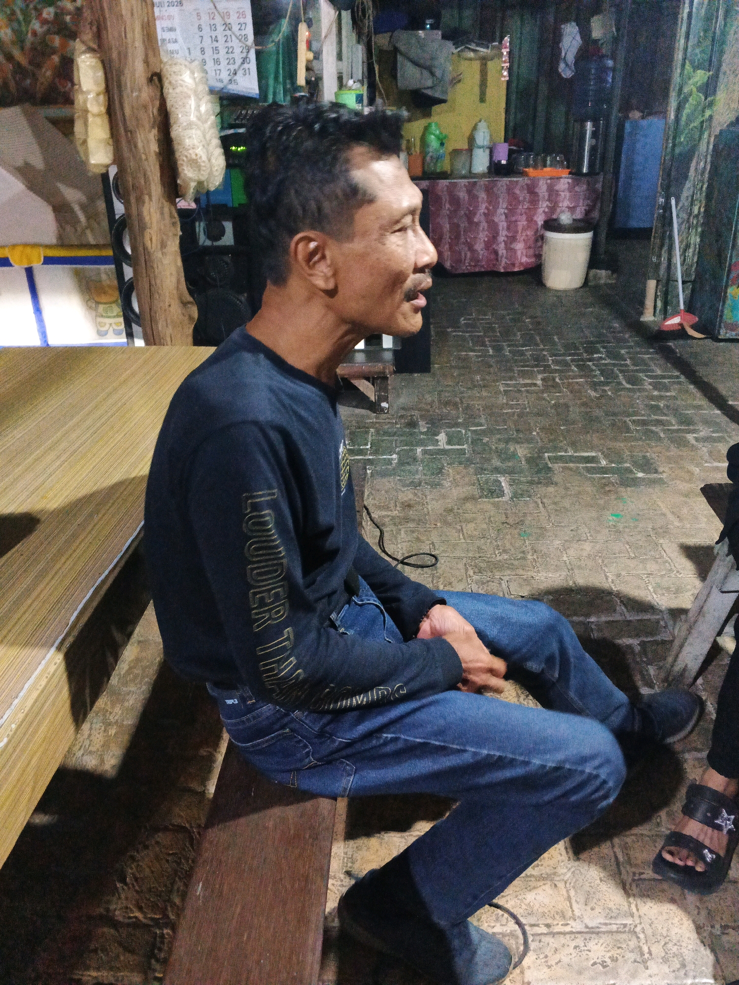 In Ungaran, Indonesia, a man with a mustache and short, dark hair is sitting on a wooden bench. He is wearing a dark blue long-sleeved shirt with gold lettering on the sleeve that appears to read "LOUDER TH". His jeans are also dark blue. He is sitting with his legs crossed and his hands clasped in his lap. The bench is made of wood and has a light-colored, wood-grain patterned surface. In the background, there is a brick floor, a table covered with a pink and white patterned cloth, and shelves with various items, including bottles and glasses. A water dispenser is visible on one of the shelves. There are also some items hanging on a wooden post in the foreground, including what appears to be packaged food. The scene appears to be an outdoor or semi-outdoor area.
