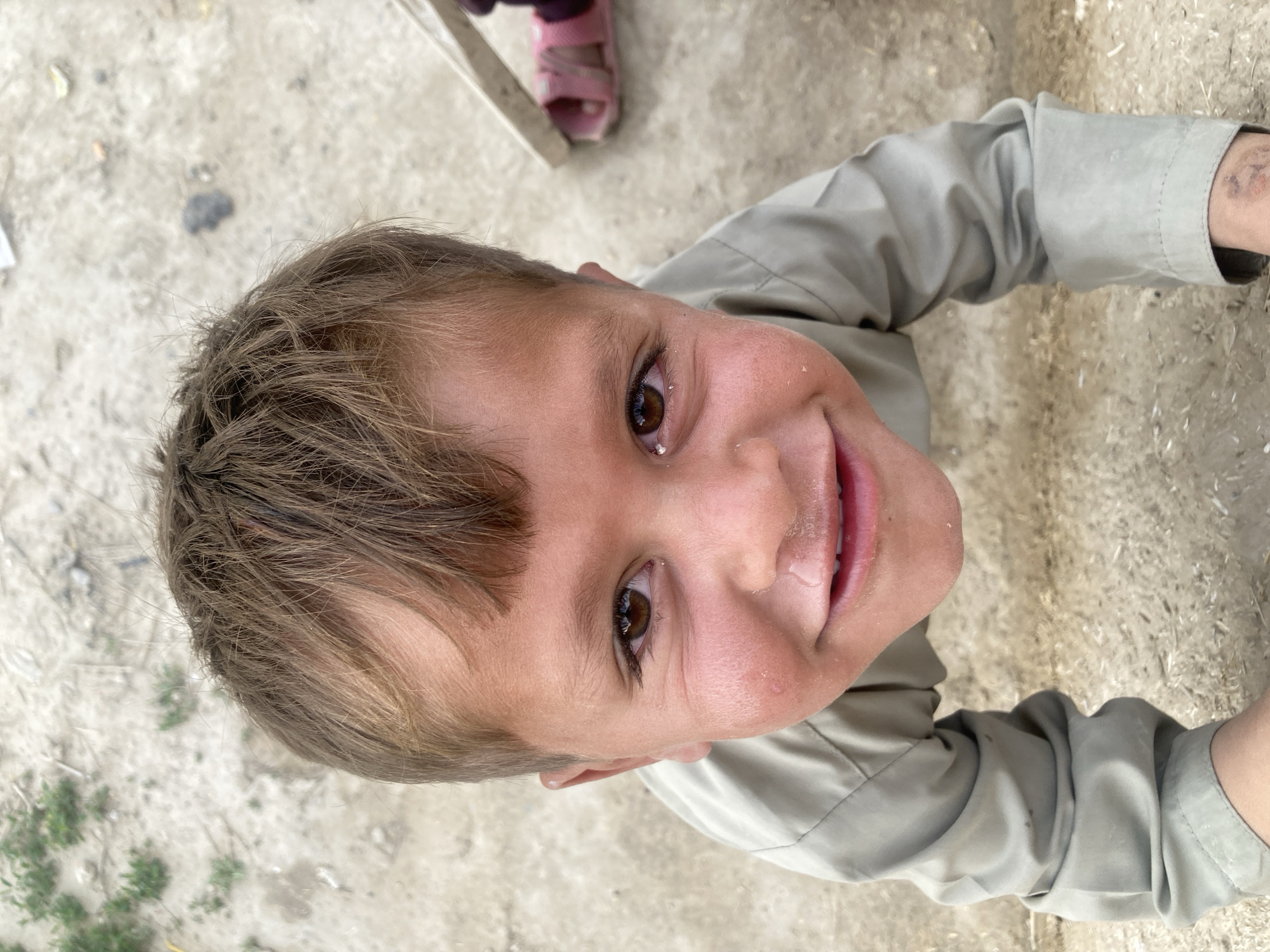 A close-up, high-angle portrait shot depicts a young child, approximately 3-6 years old, situated outdoors on dusty, light-brown ground with sparse green vegetation, within the geographical context of Quetta City Tehsil, Pakistan. The child, possessing medium-brown hair and brown eyes, gazes directly upward towards the camera with a wide smile, revealing upper teeth. Trace amounts of clear liquid are visible around both eyes and on the upper lip/philtrum region. A minor reddish mark is present on the child's left cheek. The child wears a light-beige or light-gray long-sleeved garment, likely a traditional shirt, with discernible fabric folds on the sleeves. The child's posture indicates a low position, possibly kneeling or bent forward, with both arms extending out of the bottom of the frame. In the upper right background, out of focus, a portion of a pink sandal and a light-colored wooden element are partially visible on the ground.