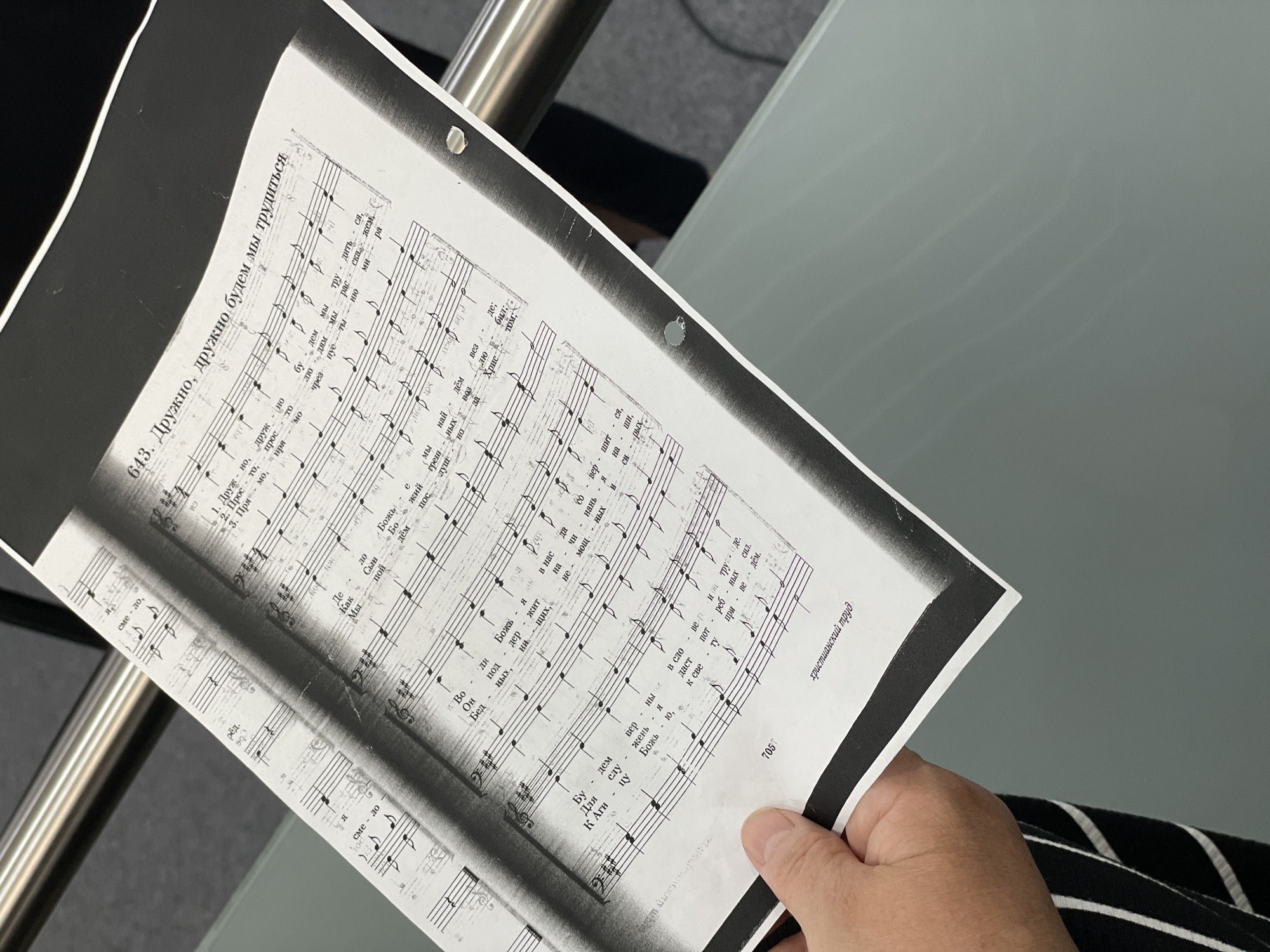 This is a close-up, slightly angled shot of a person holding a sheet of music. The sheet music is black and white and appears to be a scanned or printed document. It contains musical notation with lyrics written above the notes in Cyrillic script. The title of the piece is visible at the top of the page: "643. Дружно, дружно будем мы трудиться." (643. Together, together we will work.) The page number "705" is visible at the bottom left, and what appears to be a publication title "христианский труд" (Christian labor) is at the bottom right.
The person's hand is visible in the foreground, holding the bottom left corner of the music sheet. They are wearing a black and white striped garment. The background is blurred but shows a metallic railing and a grey surface, possibly a table or desk. The lighting suggests an indoor environment. The scene is forensic in its detail, capturing the physical object of the music sheet and the immediate context of its handling. No specific location in Lich, Germany can be determined from the provided image.