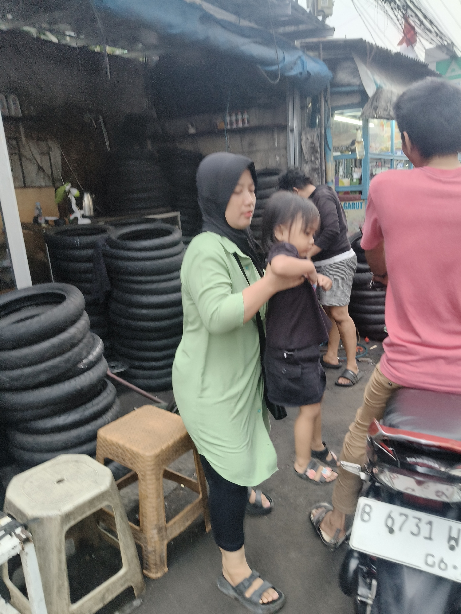 The image depicts an outdoor scene in South Tangerang, Indonesia, likely a street-side establishment selling motorcycle tires. In the foreground, a woman wearing a light green hijab and tunic is holding a young child. The child is dressed in a black t-shirt and shorts. To the right of the woman, a person in a pink shirt is seated on a motorcycle. The background is filled with stacks of black motorcycle tires. Two plastic stools, one light gray and one brown, are placed near the woman. The ground appears to be paved. Several other individuals are visible in the background, some near a stall. The overall environment suggests a commercial area, possibly a market or roadside shop.