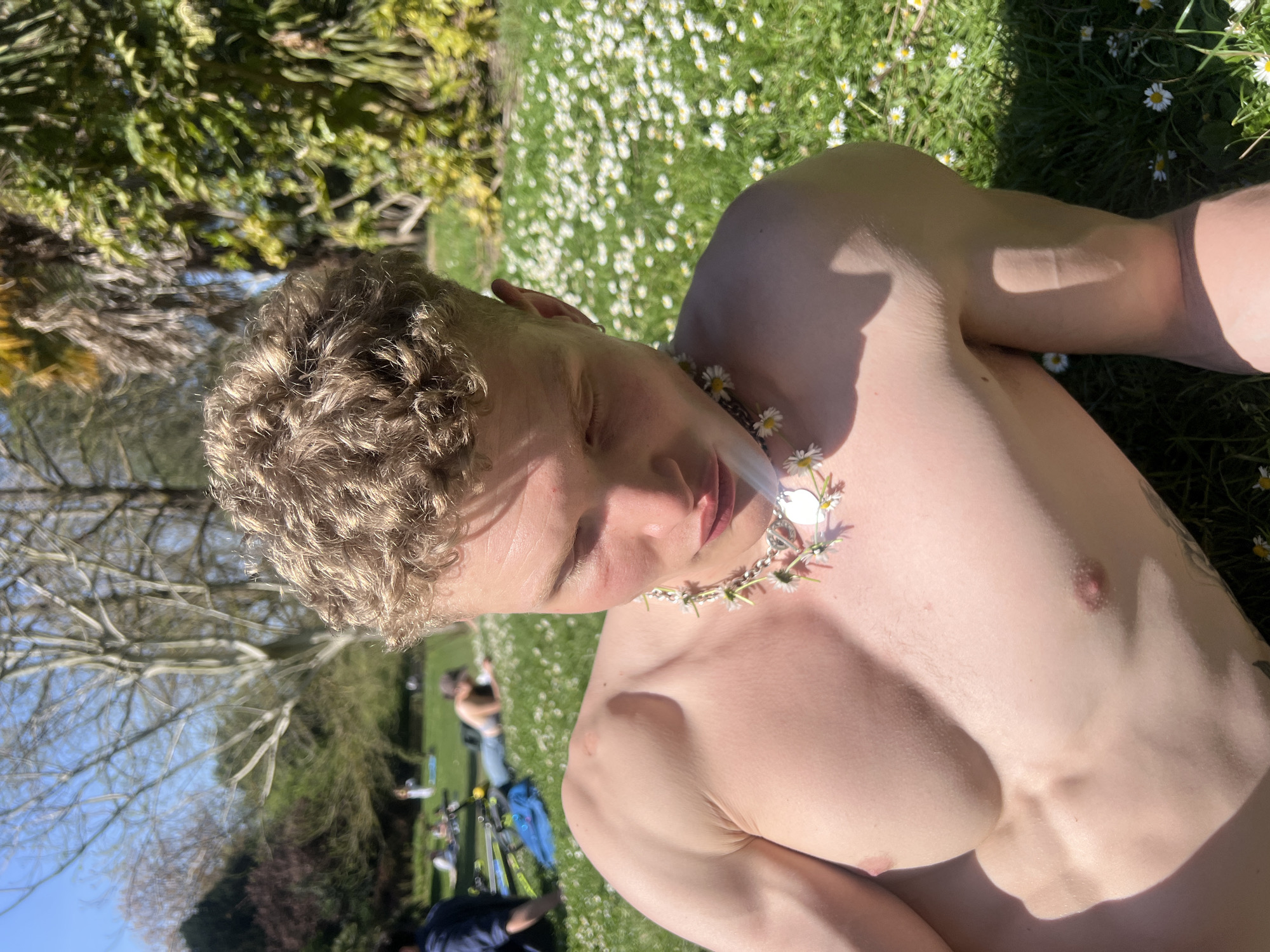 The image shows a bare-chested individual with curly blonde hair lying in a grassy field. The person is wearing a daisy chain necklace. The background includes a field of daisies, trees, and other people in the distance, with bicycles visible as well. This scene appears to be taking place in a park, possibly in London, United Kingdom.