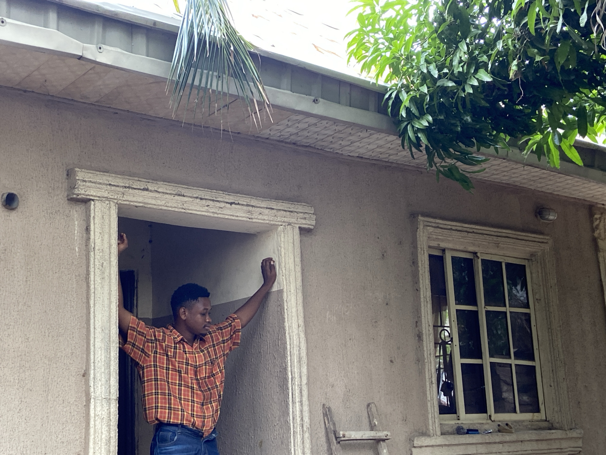 A man in a plaid shirt stands in a doorway of a stucco building. The man is dark-skinned and has short black hair. He is wearing blue jeans and has his arms raised, touching the doorframe. To the right of the doorway is a window with a white frame and panes. Underneath the window are a screwdriver, a blue object, and a pair of sandals. A ladder is partially visible near the doorway. Green foliage hangs over the right side of the building's roof, and a palm frond hangs over the left. The building appears to be in Mararaba, Nigeria.