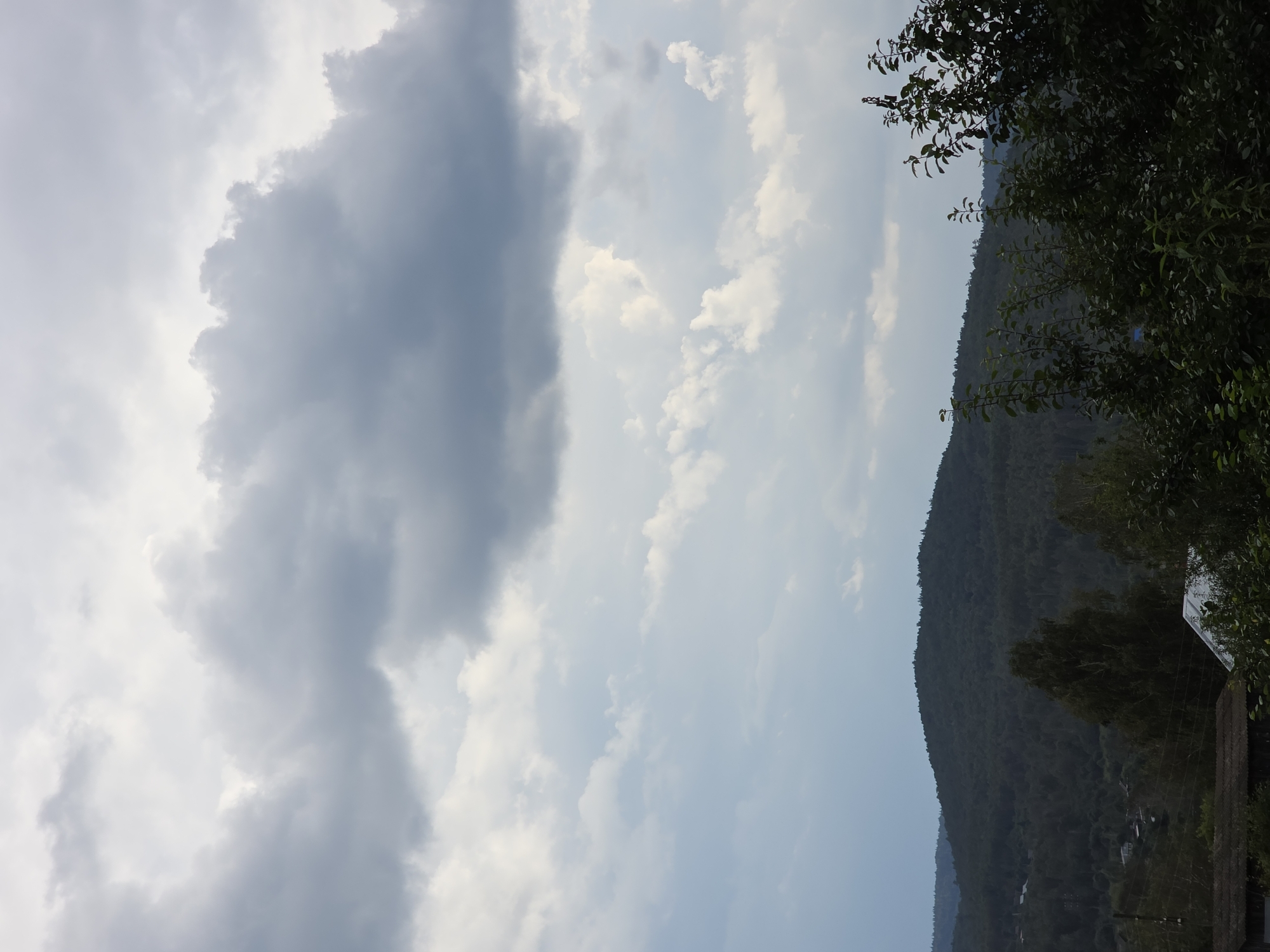 The image depicts a landscape dominated by a vast expanse of cloudy sky, with a large, dark grey cloud mass situated in the upper portion of the frame. Below the sky, rolling, densely forested hills recede into the distance. In the foreground, the tops of various trees and some rooftops of rural buildings are visible. The overall atmosphere is subdued, suggesting an overcast or perhaps pre-storm condition. The scene is consistent with the natural environment found in and around Magnitka, Russia, a region characterized by its hilly terrain and extensive forests. There are no people or discernible actions captured in the image.