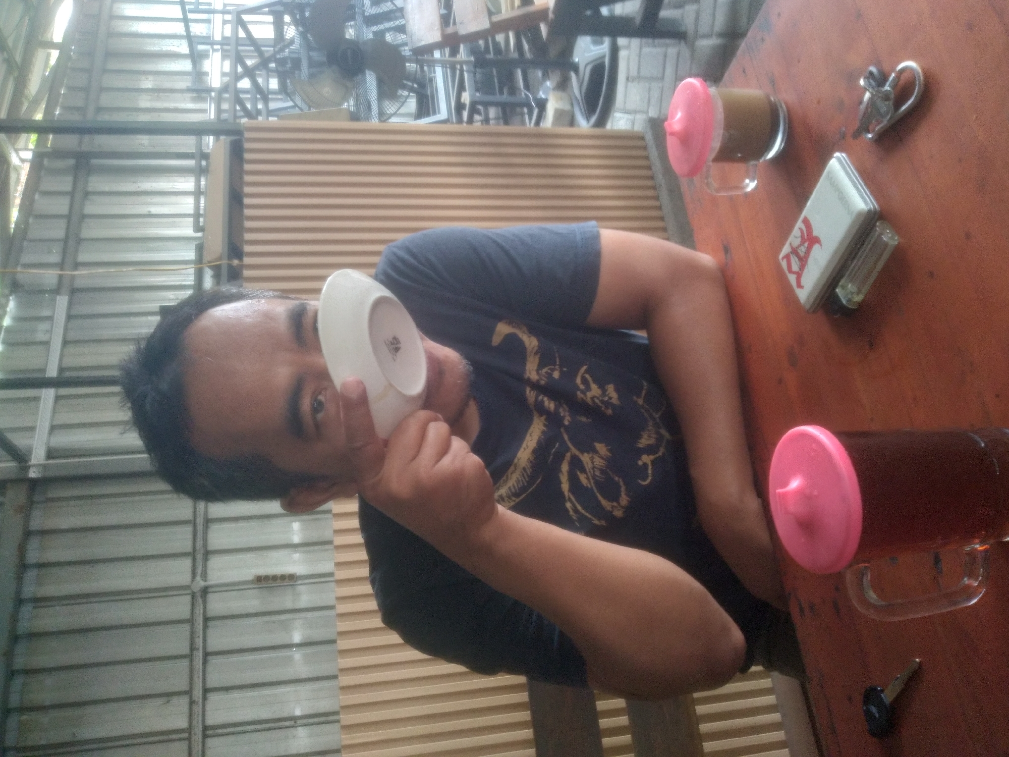 An outdoor cafe scene, location unknown, features a man sitting at a wooden table. The man, with short dark hair and a goatee, is holding a white cup to his mouth. He is wearing a dark blue t-shirt with a graphic design. On the table, there are two mugs with pink lids, one containing a dark liquid and the other a lighter brown beverage. A lighter, a pack of cigarettes, and a key are also present on the table. The background consists of a corrugated metal wall and wooden slats. A standing fan is visible in the distance. The lighting suggests it is daytime, possibly overcast.