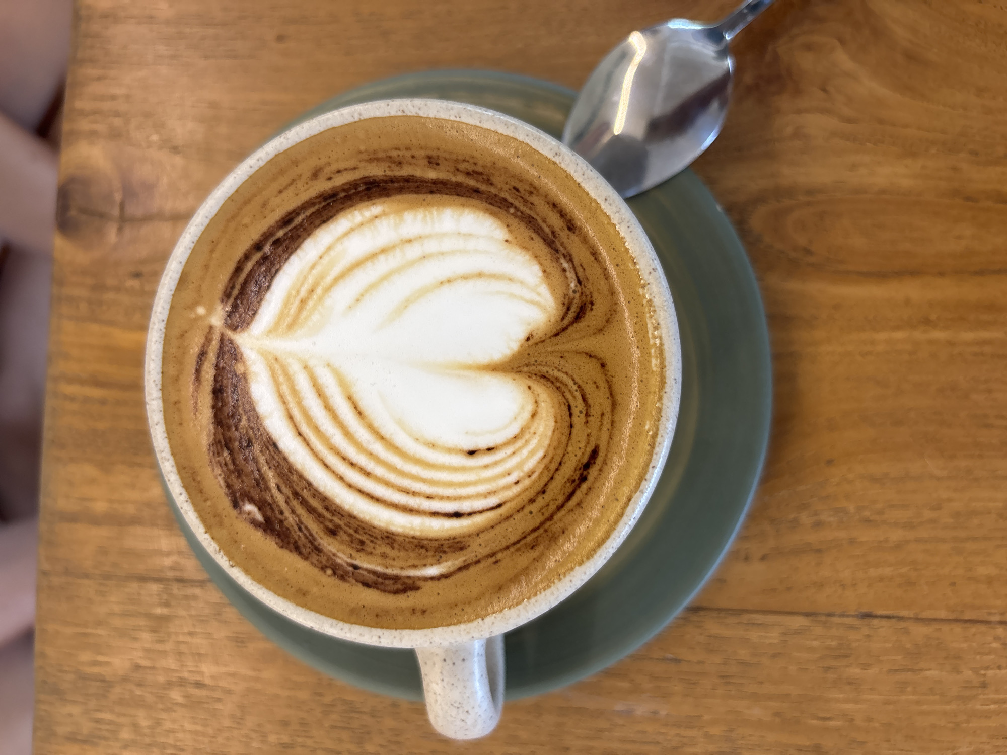 A close-up, overhead shot shows a cup of latte art on a wooden table. The latte art features a white, heart-shaped design with concentric brown rings radiating outwards, creating a detailed pattern. The cup is speckled white with a handle and rests on a green saucer. A silver spoon lies to the right of the saucer, its reflection visible on the curved surface. The background is a warm, wooden tabletop with visible grain patterns. The scene appears to be in Kuta Selatan, Indonesia, possibly at a cafe or restaurant given the presentation of the coffee.