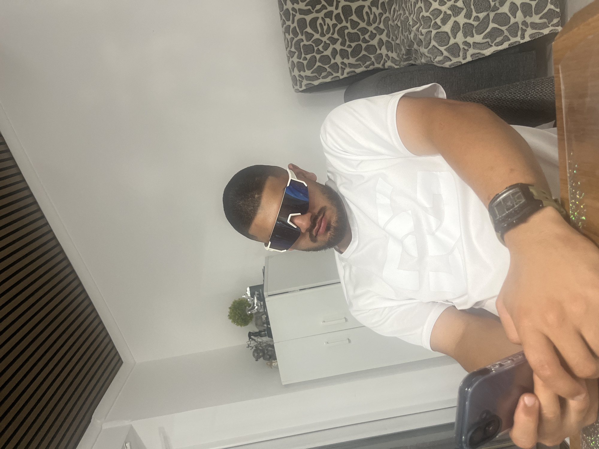In an indoor setting, a man wearing a white t-shirt and large white-framed sunglasses is seated at a wooden table. He is holding a smartphone in his left hand, with his right hand resting on the table. A dark-colored wristwatch is visible on his right wrist. Behind him, a patterned sofa is visible. To his left, a white cabinet with two doors is present, topped with decorative items including figurines and a small plant. The ceiling features a striped design. The location is identified as Rosenovo, Bulgaria.
