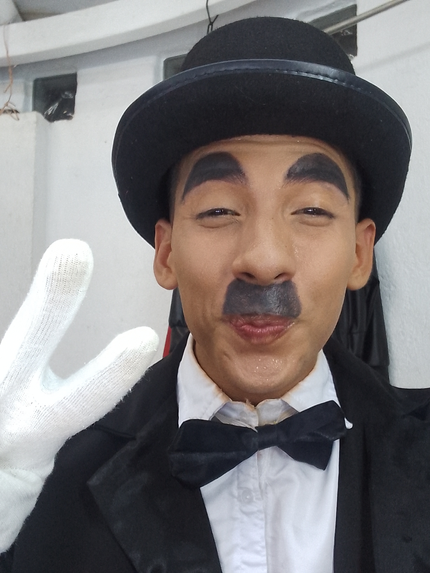 A person dressed as Charlie Chaplin poses indoors. The person wears a black bowler hat, black painted eyebrows and moustache, a white collared shirt with a black bow tie, a black suit jacket, and a white glove on their left hand, which is raised in a peace sign. The individual has a sweaty complexion. The background appears to be a white wall with a small, dark vent or opening near the top left. A clothes hanger with what looks like a dark garment is partially visible behind the person, on the right side of the image. Location details such as the specific city in Venezuela are not readily discernible from the visual information provided in the image.