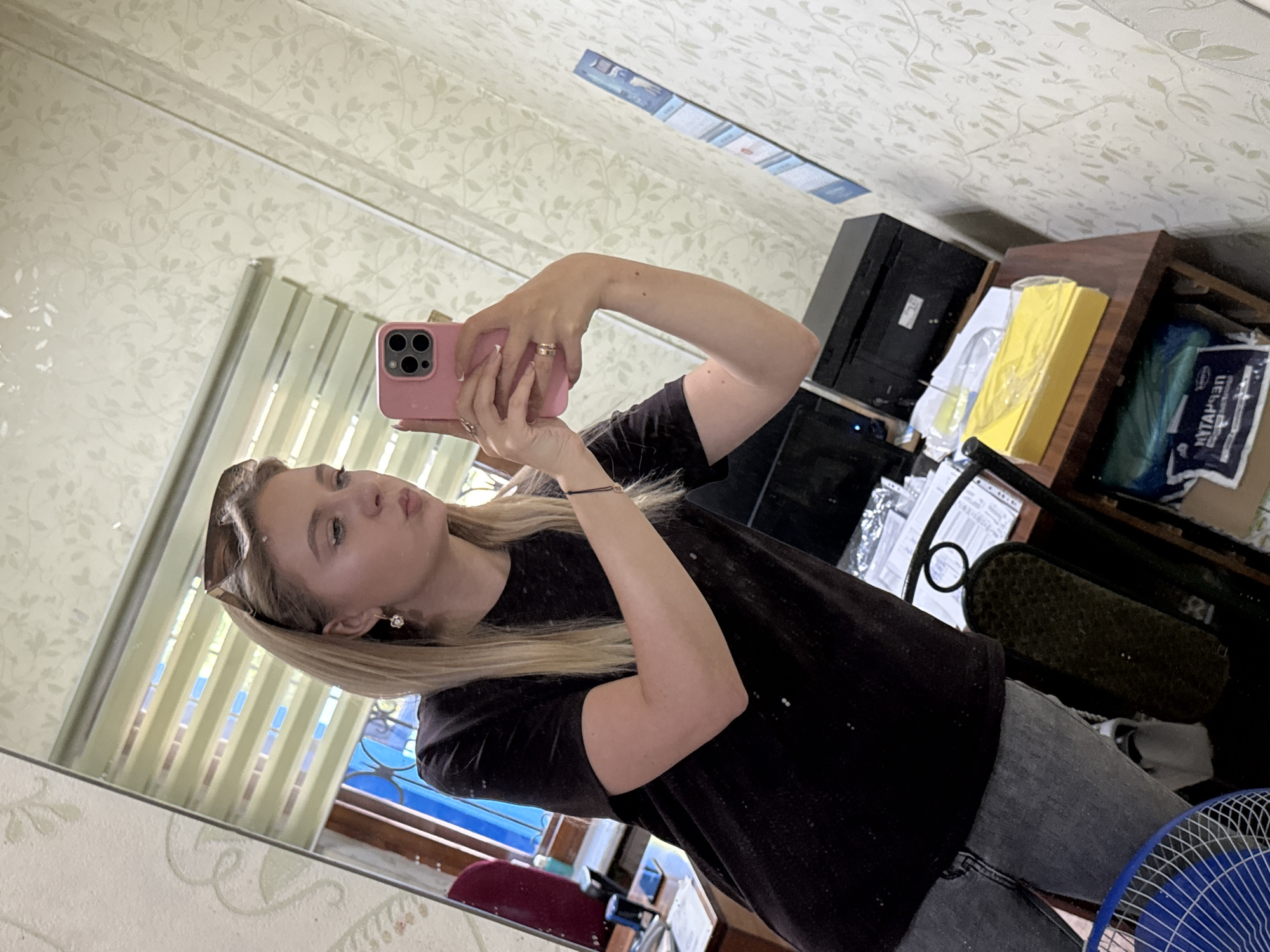 A woman with blonde hair and sunglasses is taking a selfie in a mirror. She is wearing a black t-shirt and gray jeans. She is holding a pink smartphone in her right hand and her left hand is resting on her hip. In the background, there is a desk with a printer and some papers. The room has patterned wallpaper and blinds on the window.