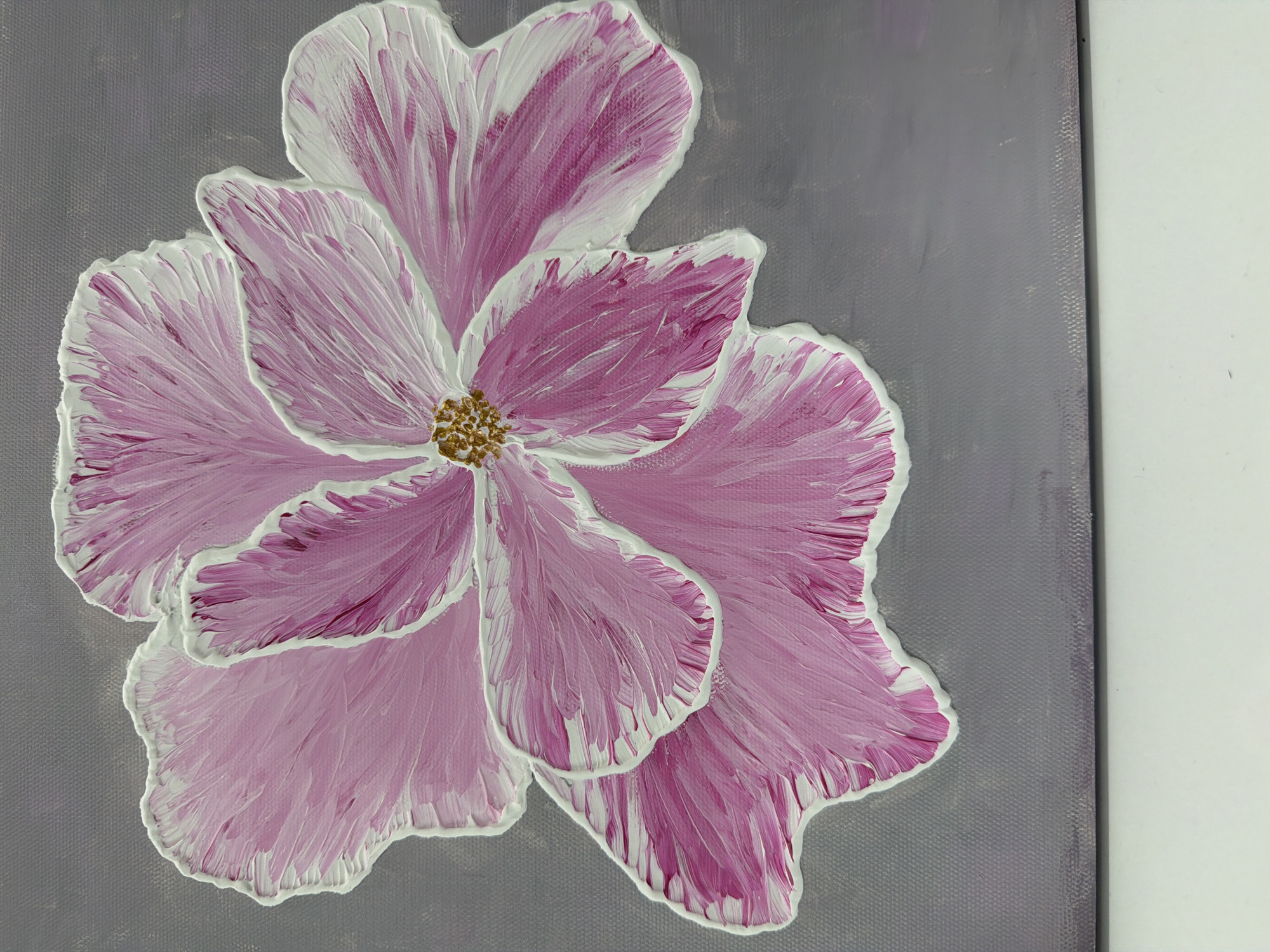 The image is a close-up, high-angle shot of an acrylic painting of a pink flower with white-edged petals against a grey background. The flower is the central subject, filling most of the frame. The petals are rendered with thick, textured brushstrokes, showing variations of pink and white. The edges of the petals are heavily textured with white paint, giving them a raised, sculpted appearance. The center of the flower features a cluster of golden-brown stamen. The grey background appears to be canvas texture, with some subtle variations in tone. There are no people, actions, interactions, or environmental elements outside of the painting itself. The scene is indoors, with a white surface visible at the bottom of the frame, suggesting the painting is resting on a table or shelf. The location in the city of Bac Nha Trang Ward, Vietnam, is not discernible from the image.