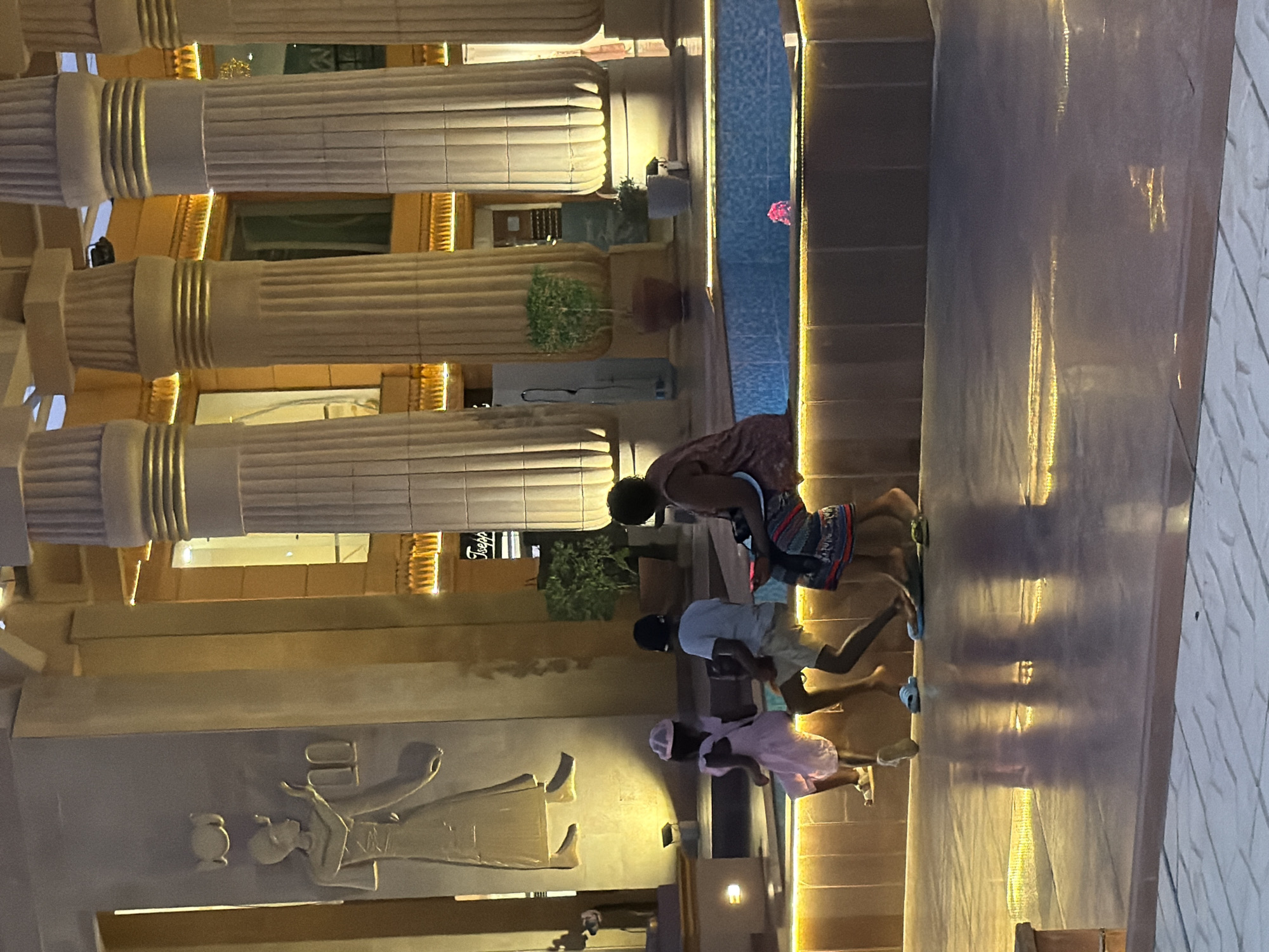 The image depicts an outdoor scene at dusk, possibly in a resort or entertainment complex in Sharm El Sheikh, Egypt. In the foreground, three children and an adult are seated on the edge of a decorative water feature or pool. The water is illuminated with pink lighting, and a single pink flower floats on its surface. The children are casually dressed, with one wearing a pink dress and hat, another in a white t-shirt and shorts, and the third in a t-shirt and patterned shorts. The adult appears to be wearing a dark top and a patterned skirt.
The background features architecture inspired by ancient Egyptian styles, with massive fluted columns that are illuminated from below, creating a warm glow. A large relief carving of a figure in ancient Egyptian attire is visible on a pillar to the left. Potted plants are strategically placed around the architectural elements, adding a touch of greenery. The ground in the foreground is reflective, showing the illuminated columns and the figures, suggesting it is a polished surface such as marble or polished concrete. The overall ambiance is that of a well-lit public space designed for leisure and entertainment, with a clear nod to Egyptian heritage.