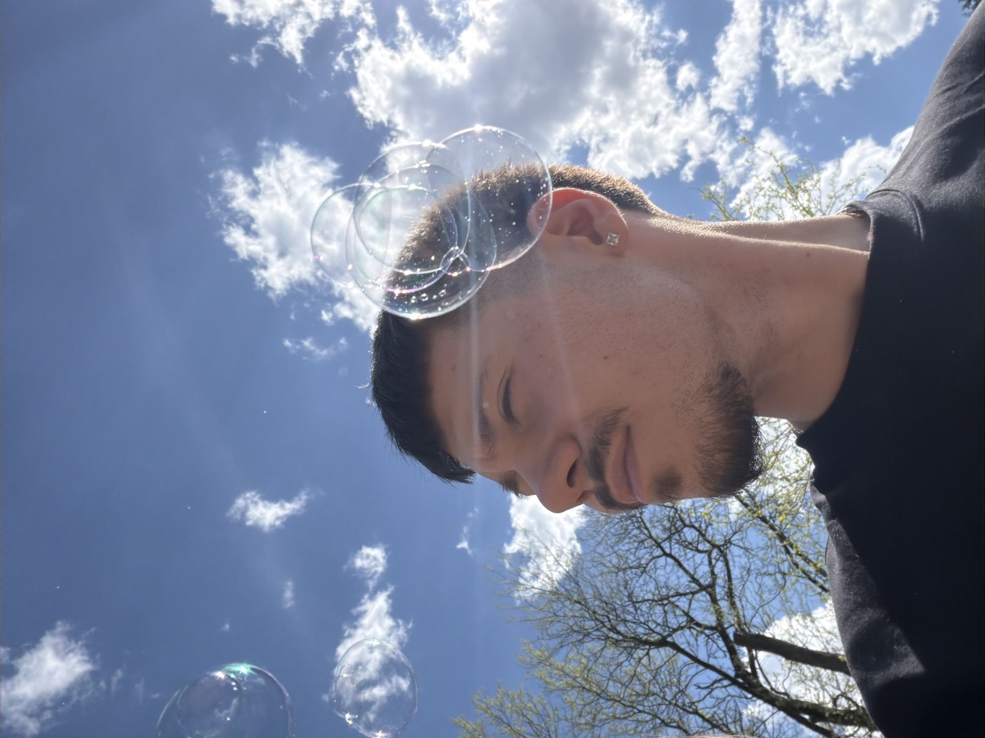 A low-angle shot shows a man with short dark hair and a neatly trimmed beard. He has light skin and wears a small stud earring. He looks downward with his eyes partially closed. He is wearing a black shirt or jacket. Soap bubbles float near his head and in the bottom left of the frame. Behind him is a blue sky with puffy white clouds. Bare tree branches are visible in the lower right, and the image appears to have been taken outdoors, possibly in Sofia, Bulgaria, given the request.