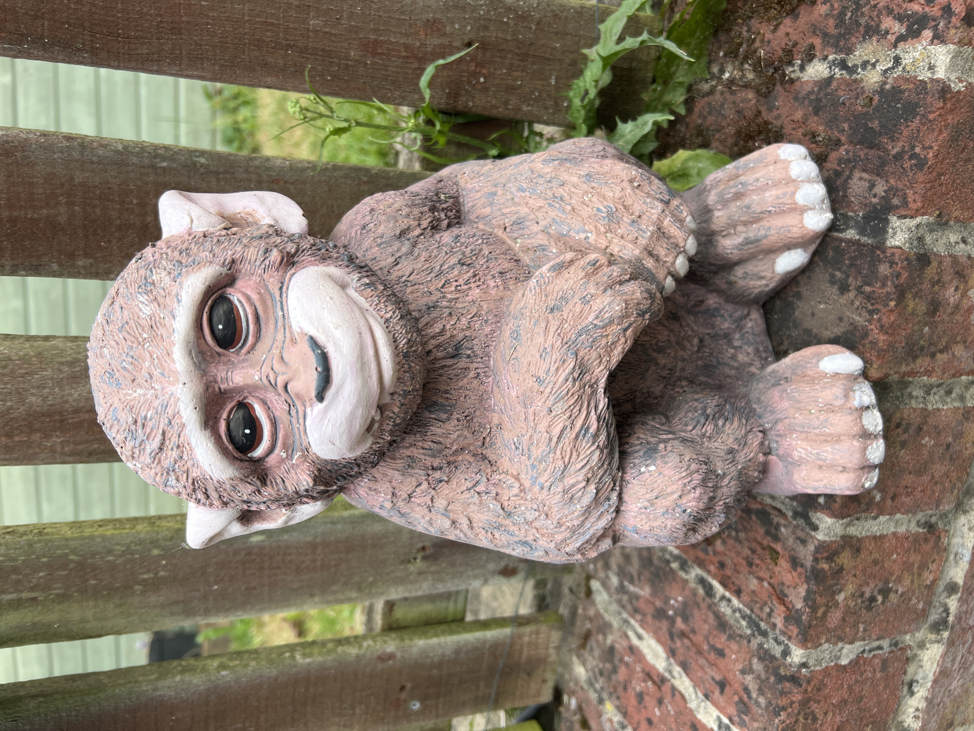 A garden gnome statue of a stylized monkey is resting on a corner where a wooden fence meets a brick wall. The monkey is light brown with gray patches and white accents on its face, ears, and the tips of its toes. Its legs are crossed and it appears to be leaning back slightly. The wooden fence has a weathered look and the brick wall shows signs of age. Green foliage is visible behind the fence and around the base of the wall. The photo was taken in the United Kingdom, in the city of Basingstoke and Deane.
