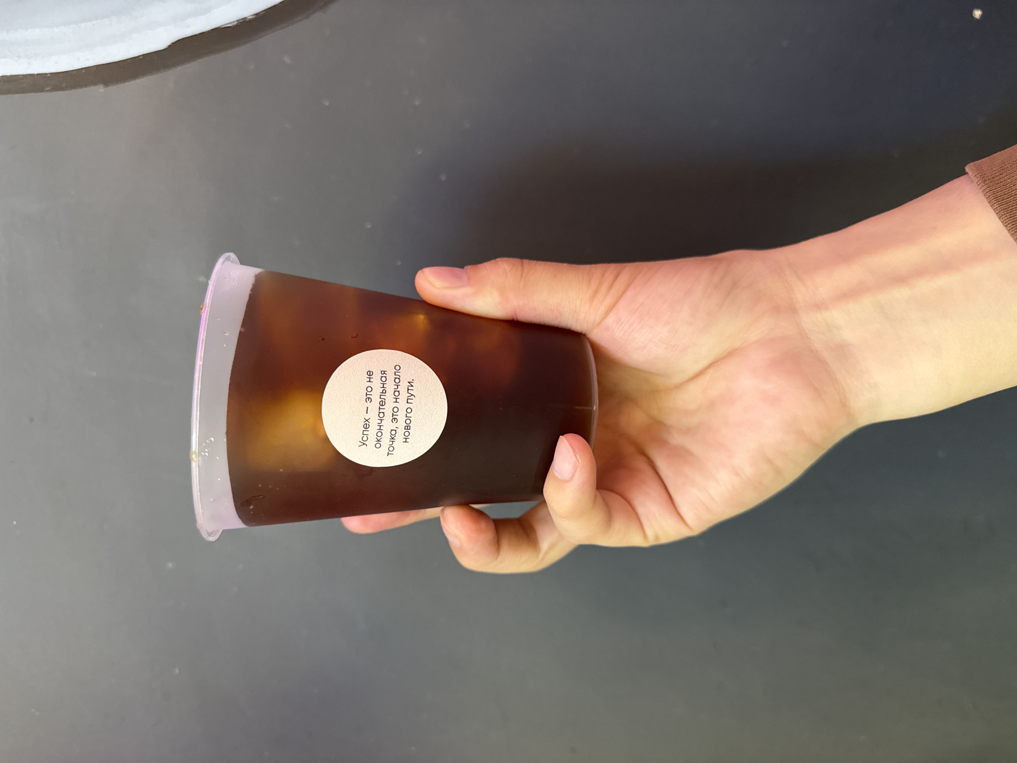A person's hand holds a clear plastic cup filled with a dark liquid and ice. A circular white sticker with text in Russian is affixed to the cup. The background is a dark, textured surface. The location of this scene in Veshki, Russia, is not discernible from the image.