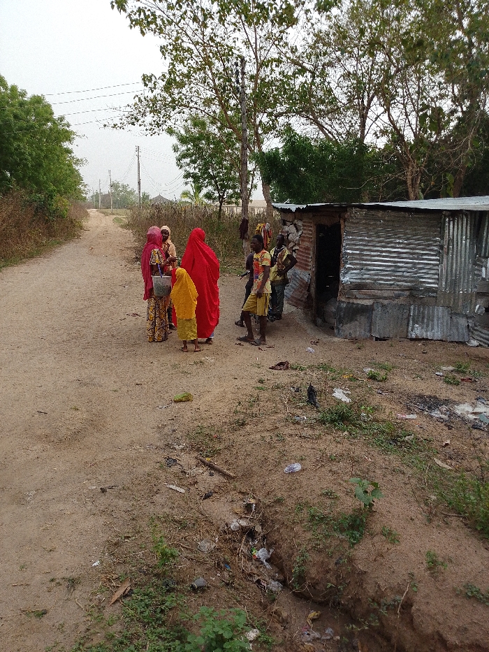 A group of people stand on a dirt road in a rural setting. Three women wear brightly colored robes, while two men stand nearby, clad in casual clothing. A small corrugated metal structure sits to the right, likely a shelter or storage space. Trash litters the ground in a nearby ditch. The scene evokes a sense of poverty and simplicity, juxtaposed with the vibrant colors of the women's attire. Overhead are utility lines, hinting at some infrastructure in the area.
