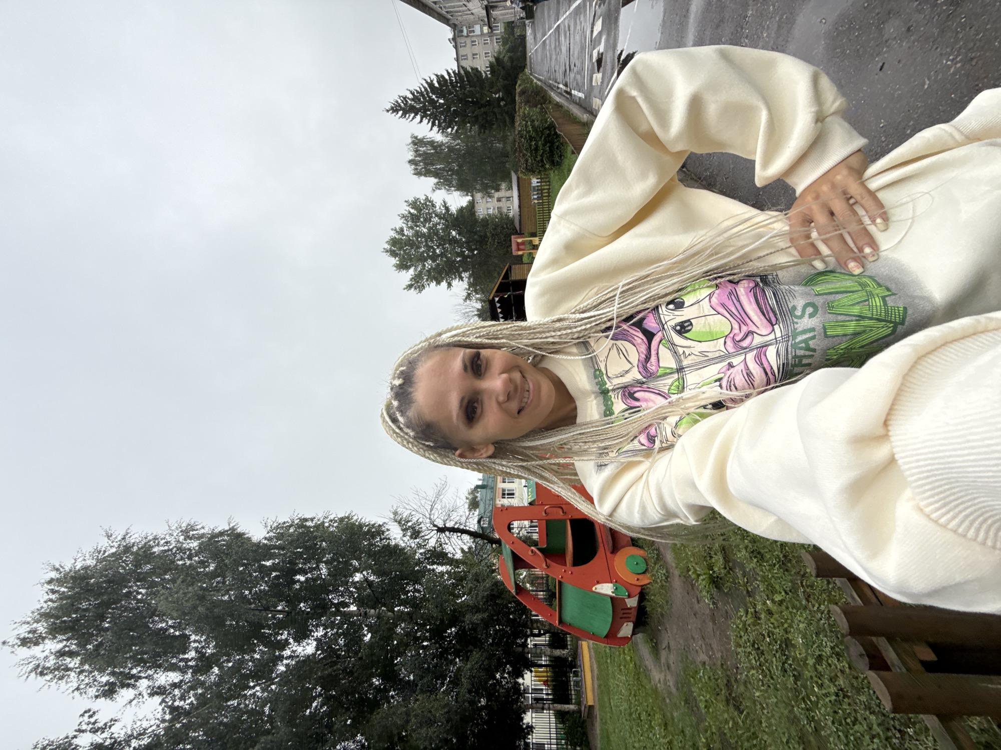 A woman with blonde braided hair is taking a selfie outdoors on a cloudy day. She is wearing a light-colored oversized sweatshirt with a graphic print. Her right hand is on her hip. Behind her, there is a playground with a red and green toy vehicle. To the right, there is a paved walkway with puddles and buildings in the background. The scene appears to be in a residential area in Rusanovo, Russia, as evidenced by the apartment buildings and typical urban playground.