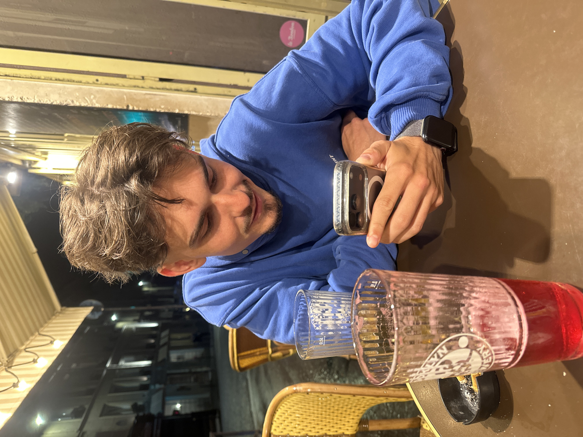 A young man with light brown hair is seated at an outdoor cafe table. He is wearing a blue sweatshirt and a smartwatch on his left wrist. He is holding a smartphone in his right hand, looking at the screen. On the table are two large glasses, one empty and one containing a red liquid. An ashtray is also present. The cafe has a wicker chair. Outside the cafe, the building is visible, with lights illuminating the scene. This photo was most likely taken in Paris.