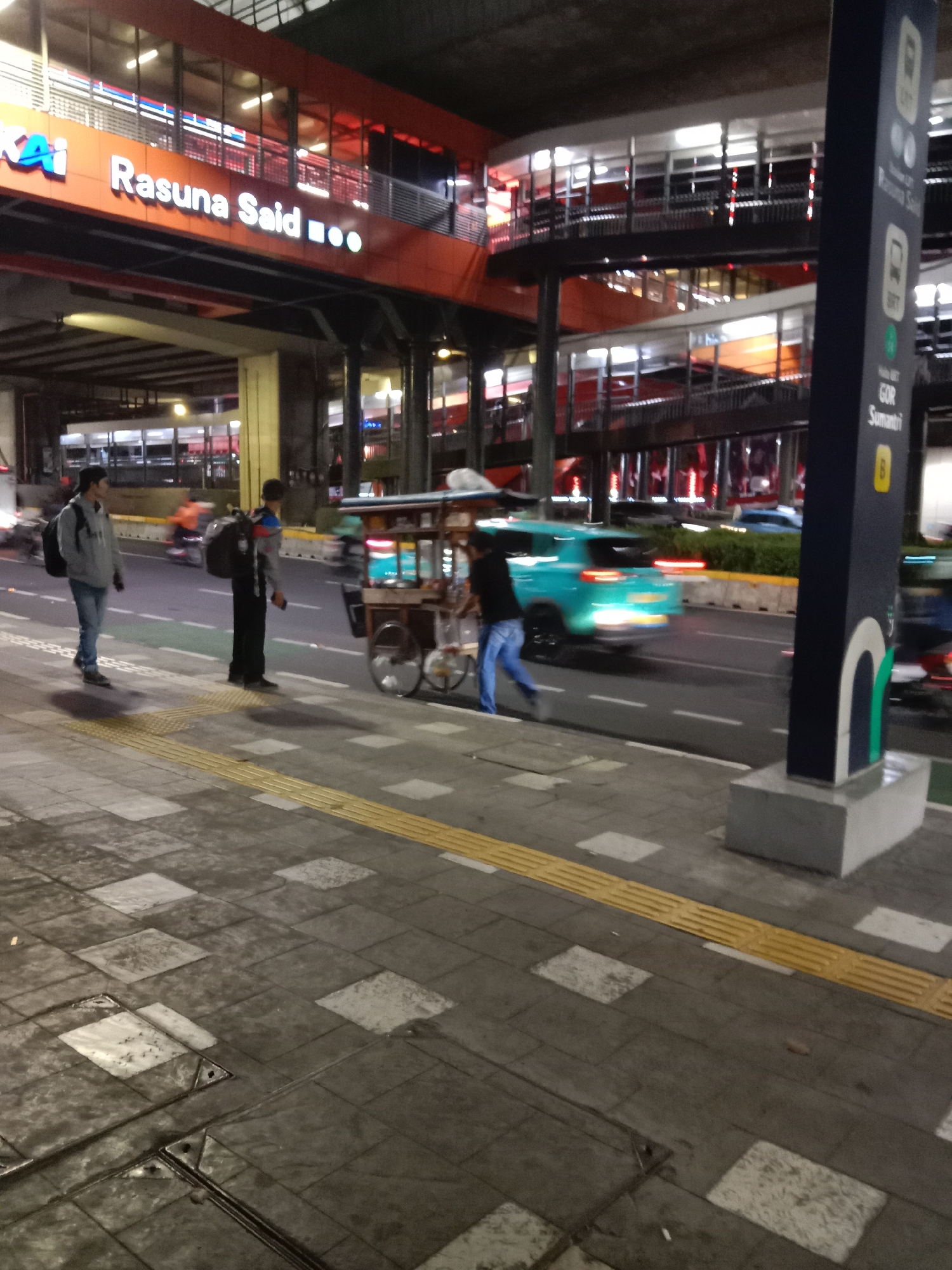 This image captures a street scene in South Jakarta, Indonesia, likely near the Rasuna Said train station, indicated by the prominent signage. In the foreground, a pedestrian walkway is paved with textured tiles, featuring a tactile paving strip for the visually impaired. On the street, two individuals are walking, one wearing a grey hoodie and backpack, the other in dark clothing with a backpack. A street vendor's cart, filled with items and equipped with wheels, is positioned on the right side of the street. A teal-colored car is in motion on the road, its taillights creating streaks of light due to the low light conditions and movement. The background reveals a multi-level transit station with orange and black architecture, illuminated by internal lighting. A tall, dark blue signpost is visible on the right, with white text and graphic icons, suggesting station information or directions. The overall atmosphere is that of a busy urban environment at dusk or night.