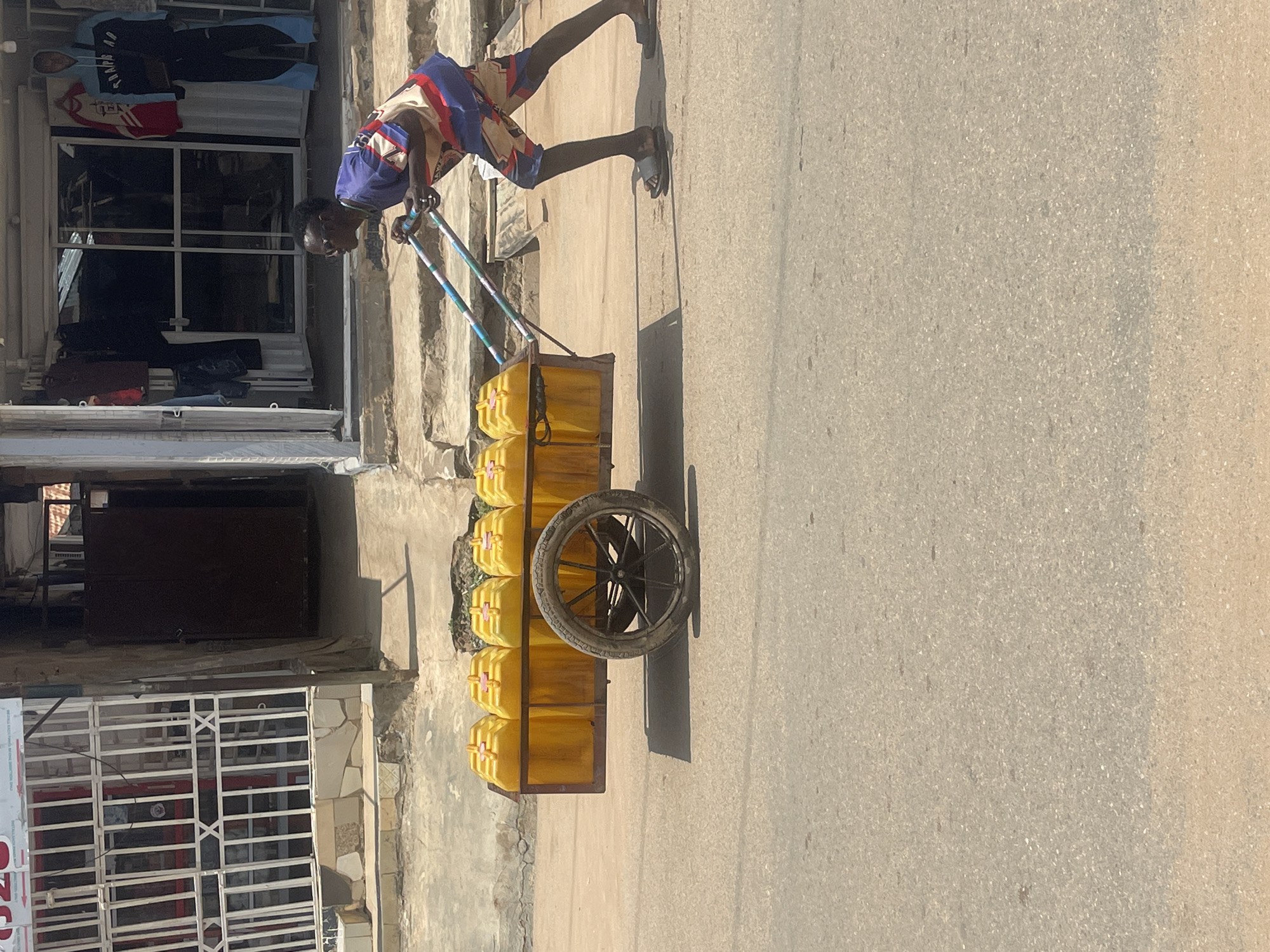 The image captures a street scene in Abuja, Nigeria. A young person, dressed in a colorful t-shirt and patterned shorts, is pushing a cart laden with numerous yellow plastic containers. The cart is a simple metal frame with a single wheel at the back. The containers are uniformly shaped and appear to be filled with liquid. The person is walking on an asphalt road, with a sidewalk and buildings visible in the background. The buildings suggest a commercial area, with shopfronts displaying various items. The overall impression is one of daily life and commerce in an urban environment.