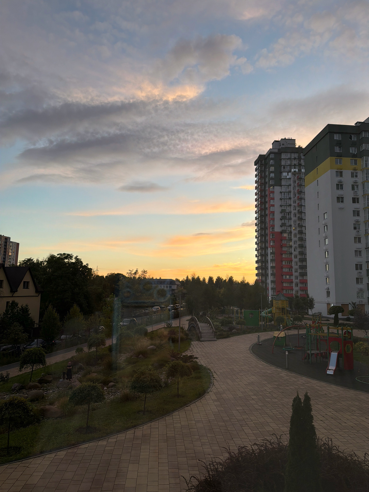 The image depicts a view of a residential complex at sunset in Kyiv, Ukraine. In the foreground, a paved pathway curves through a landscaped area with small trees and rocks. Beyond the pathway is a playground with colorful equipment. To the left, a small bridge crosses a dry creek bed, and a few people are visible in the distance within a garden area. Behind the garden, there are several residential buildings of varying heights, including a prominent multi-story building with a yellow and white facade and another tall, modern apartment building with a red accent on its side. A smaller building with a brown roof is also visible. The sky is filled with clouds tinged with the warm colors of sunset, ranging from soft orange and pink to blue and gray. The overall scene suggests a modern urban living environment with integrated green spaces and recreational facilities.