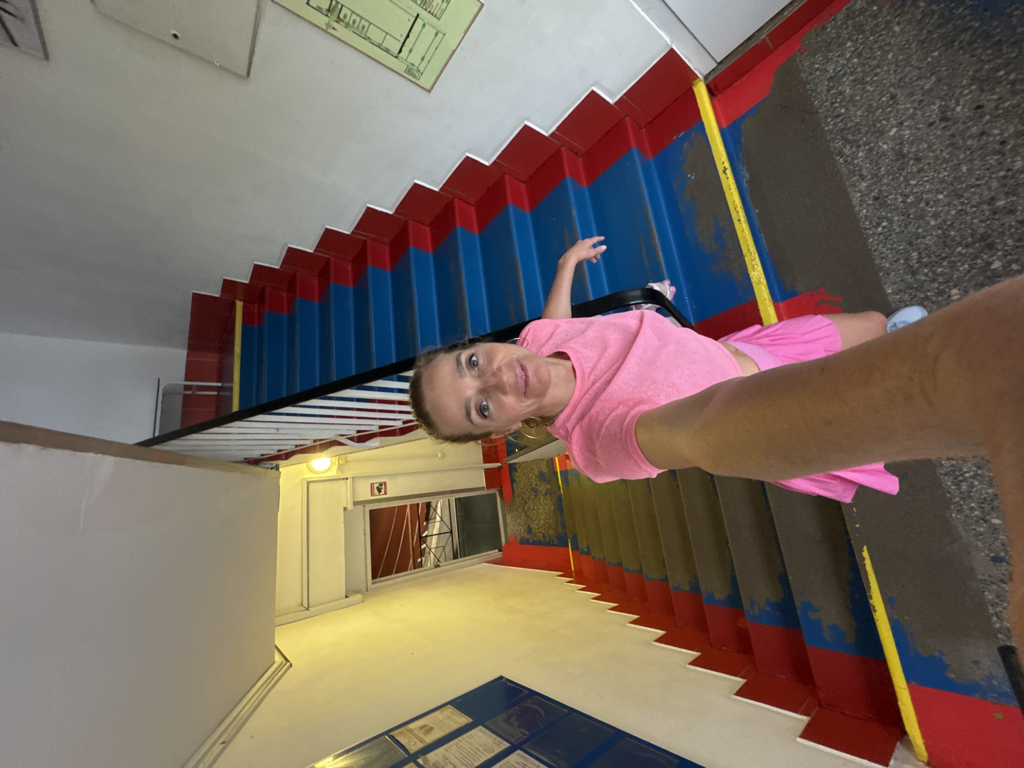 A selfie of a Caucasian woman in a pink t-shirt and pink shorts, taken inside a stairwell in Saint Petersburg, Russia. The woman is looking at the camera with a slight smile. The stairwell has steps that are painted red, blue and white. A yellow stripe lines the sides of the stairs. A metal railing runs along one side of the stairs. A diagram of the building is posted on the wall near the top of the stairs. A door and a window can be seen at the bottom of the stairs. The floor is gray.