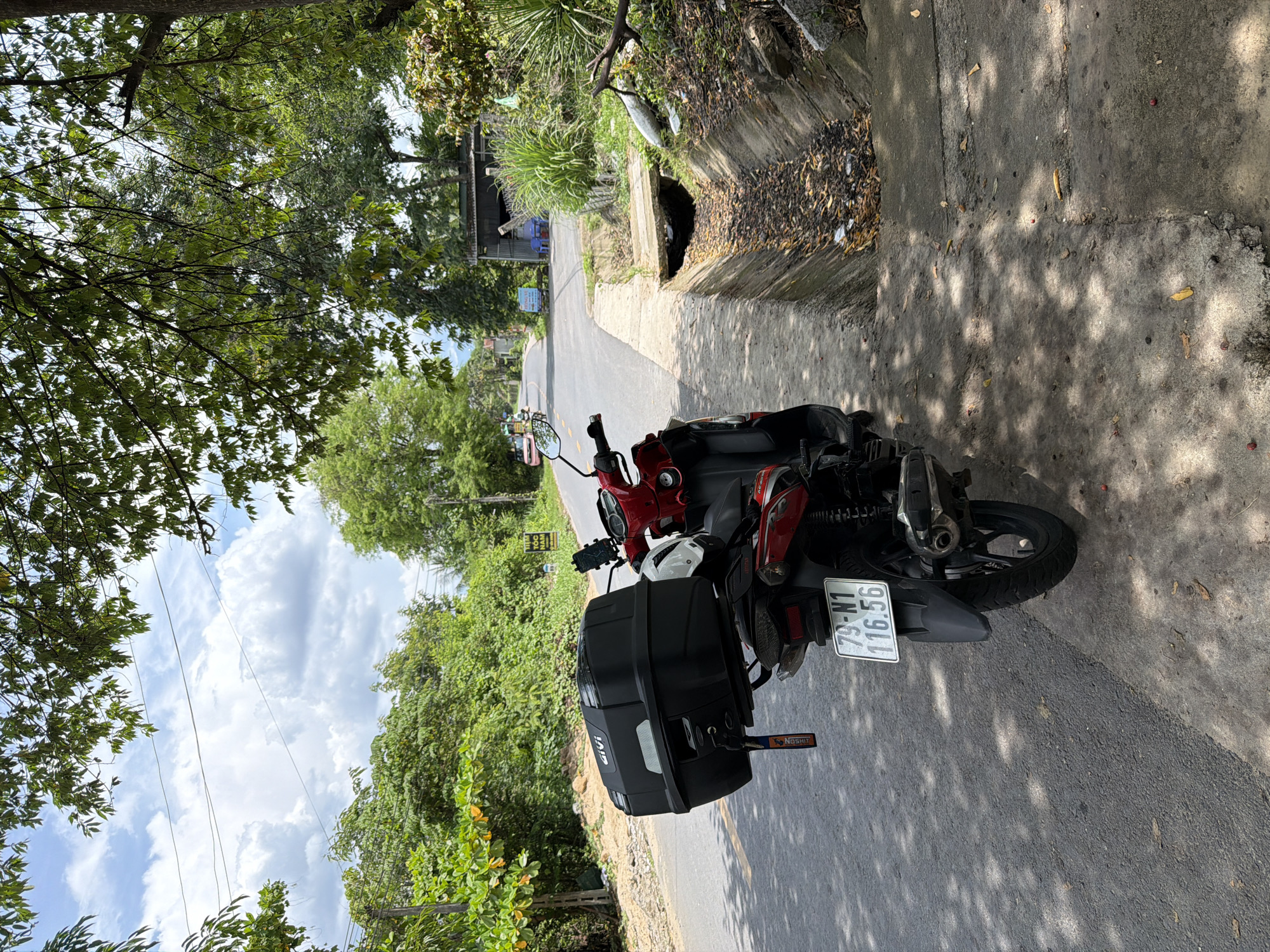 In Diên Thọ, Vietnam, a red and black scooter is parked on the right side of a paved road. The scooter faces away from the camera and is equipped with a large black GIVI top case. A white helmet rests on the seat of the scooter. The license plate of the scooter reads "79-N1 116.56". To the right of the scooter, a concrete drainage ditch runs parallel to the road, filled with debris and fallen leaves. The environment is characterized by lush green trees and vegetation lining the road, with a cloudy sky overhead. In the distance, a few buildings and other vehicles are visible along the road. The scene appears to be a roadside in a rural or semi-rural area.