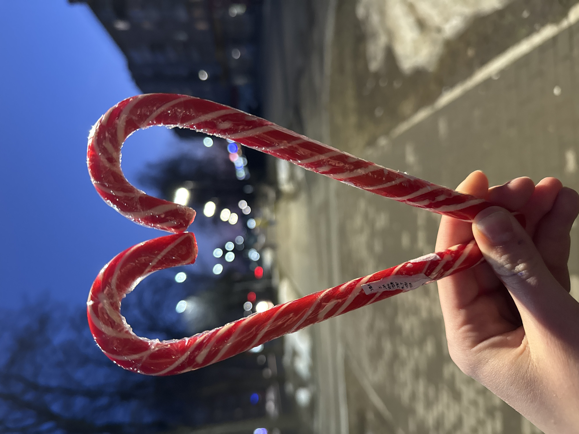 A hand holds two red and white striped candy canes in the shape of a heart against the backdrop of a blurred cityscape. The sky is a deep blue, suggesting either dusk or dawn. Out-of-focus lights, likely from street lamps or buildings, are scattered throughout the background. A building is vaguely visible in the upper left corner. The ground, partially obscured by the hand, appears to be a sidewalk or paved area. Based on the presence of Russian writing on the candy cane wrapper, the image was taken in Russia. Further detail is not resolvable.
