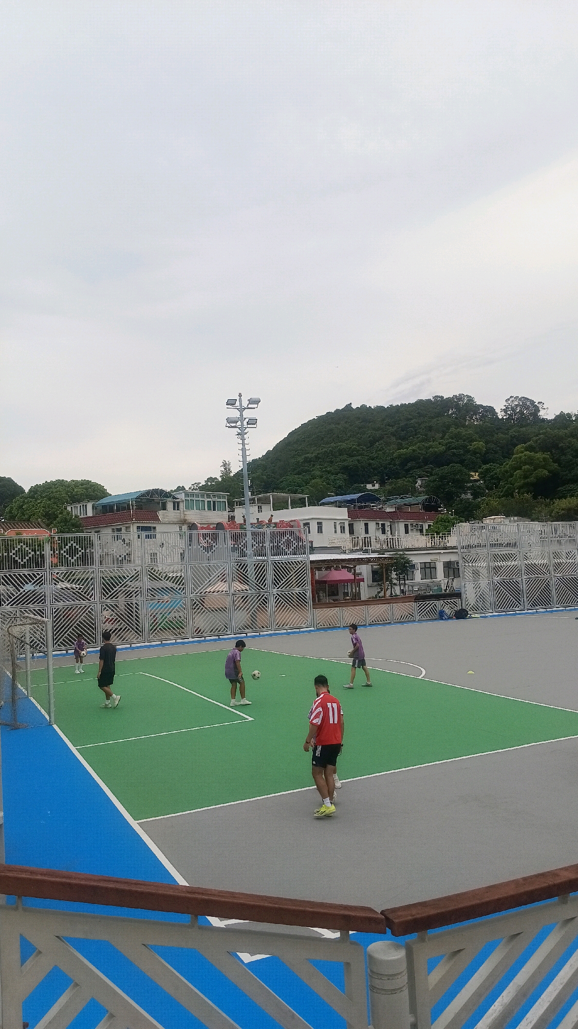 This is a vertical, eye-level, full shot of an outdoor futsal court. The court has a green playing surface with white lines marking boundaries. A blue border runs along the outside of the court. In the background, there are several buildings with white walls and red or blue roofs, and a lush green hillside covered in trees. A tall, metallic light pole with solar panels stands near the center of the court.
There are six individuals on the court, all appearing to be male teenagers or young adults, engaged in a game of futsal. One person in a red jersey with the number 11 on the back is facing away from the camera, and appears to be about to kick a ball. Another person, wearing a purple jersey, is near the center of the court with a ball at their feet. Three other individuals in purple jerseys are scattered across the court, and one person in a black shirt and shorts is standing on the left side of the court. The overall mood appears to be one of casual recreation.