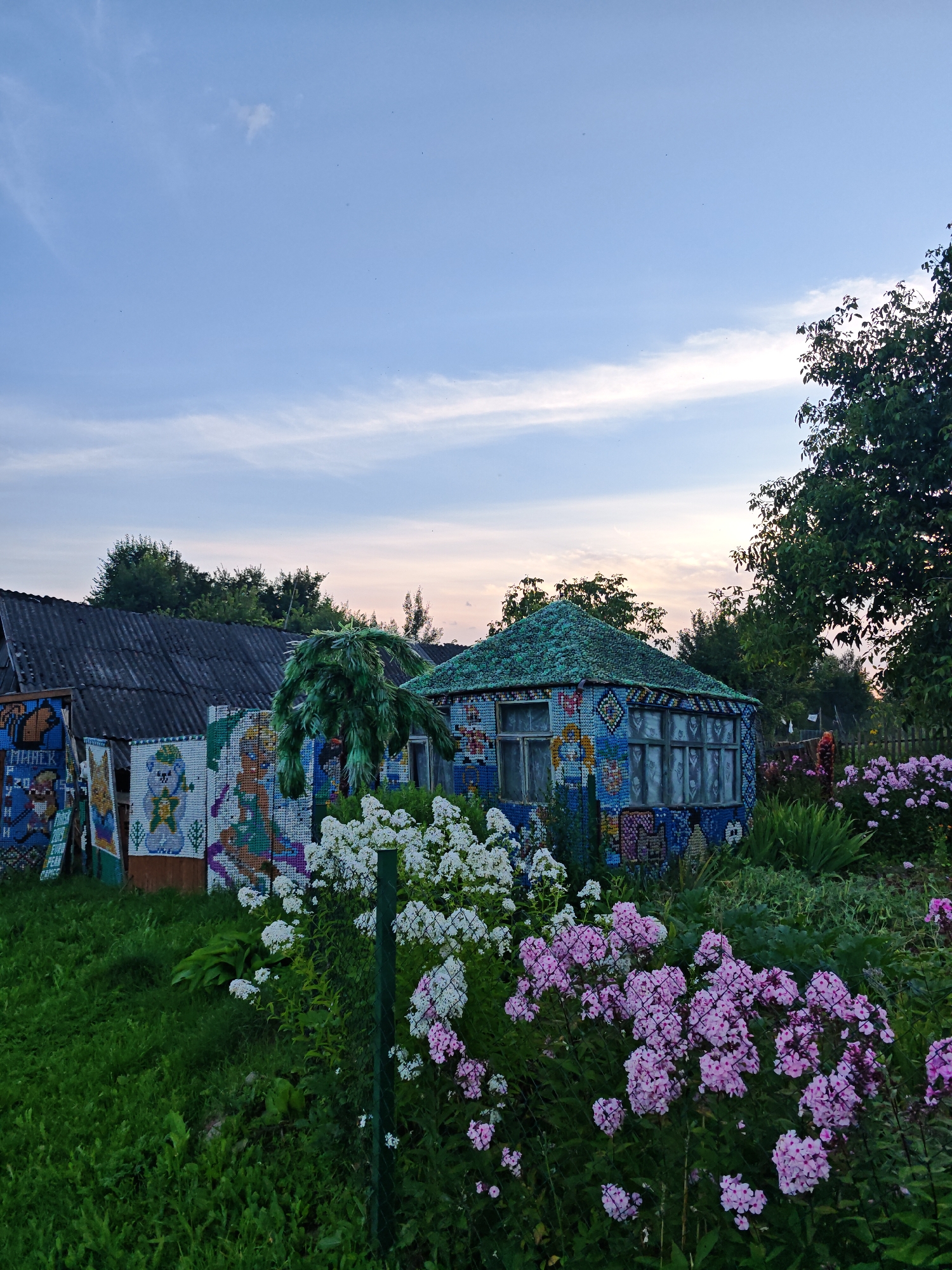 The image depicts a colorful and unusual outdoor scene in Valozhynski Sel'ski Savet, Belarus. In the foreground, a dense arrangement of pink and white phlox flowers is visible, partially obscured by a green mesh fence. Behind the flowers, a small, hexagonal building with a green, textured roof and blue, mosaic-covered walls stands prominently. The mosaics feature various colorful images and patterns. To the left of this building, another structure with a dark, corrugated roof is partially visible, also decorated with colorful mosaics, including depictions of cartoon-like characters and text. A tall, leafy green structure, resembling a stylized tree or shrub, stands between the two buildings. The background consists of lush green foliage, trees, and a pale blue sky with wispy clouds. The overall impression is of a whimsical, artistically decorated outdoor space.