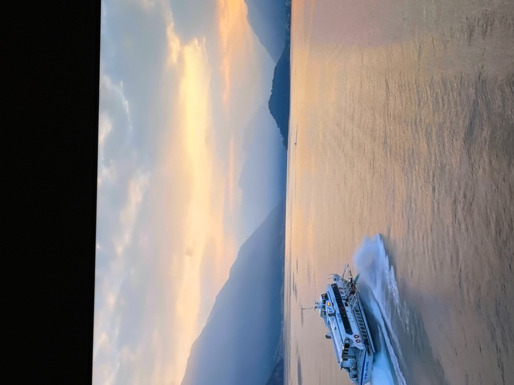 A white hydrofoil ferry, identified by the name "VOLOIRE" printed on its side, travels across a body of water. The ferry is depicted in motion, creating a significant wake behind it. In the background, majestic, mist-shrouded mountains rise from the water's edge, with a small, tree-covered island visible further out. The sky above is filled with soft, pastel clouds, illuminated by the warm hues of either sunrise or sunset, casting a golden glow on the water's surface. The overall scene conveys a sense of tranquility and natural beauty.