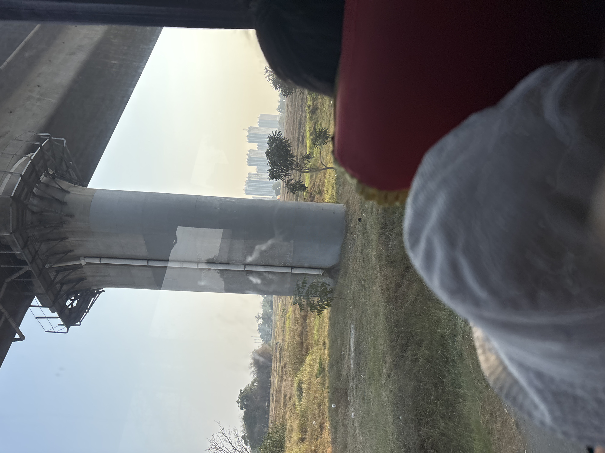 A view from inside a vehicle in Jayamukti, Indonesia, shows a massive concrete pillar supporting an elevated structure, likely part of a transportation system. The pillar dominates the left side of the frame, with pipes running vertically along its surface. Beyond the pillar, a dry, grassy field stretches towards a cluster of tall, modern buildings in the distance. Several trees are scattered across the landscape. The foreground is partially obscured by the heads of two people sitting in the vehicle, one wearing a light-colored hat and the other with dark hair visible. The overall lighting suggests it is daytime.