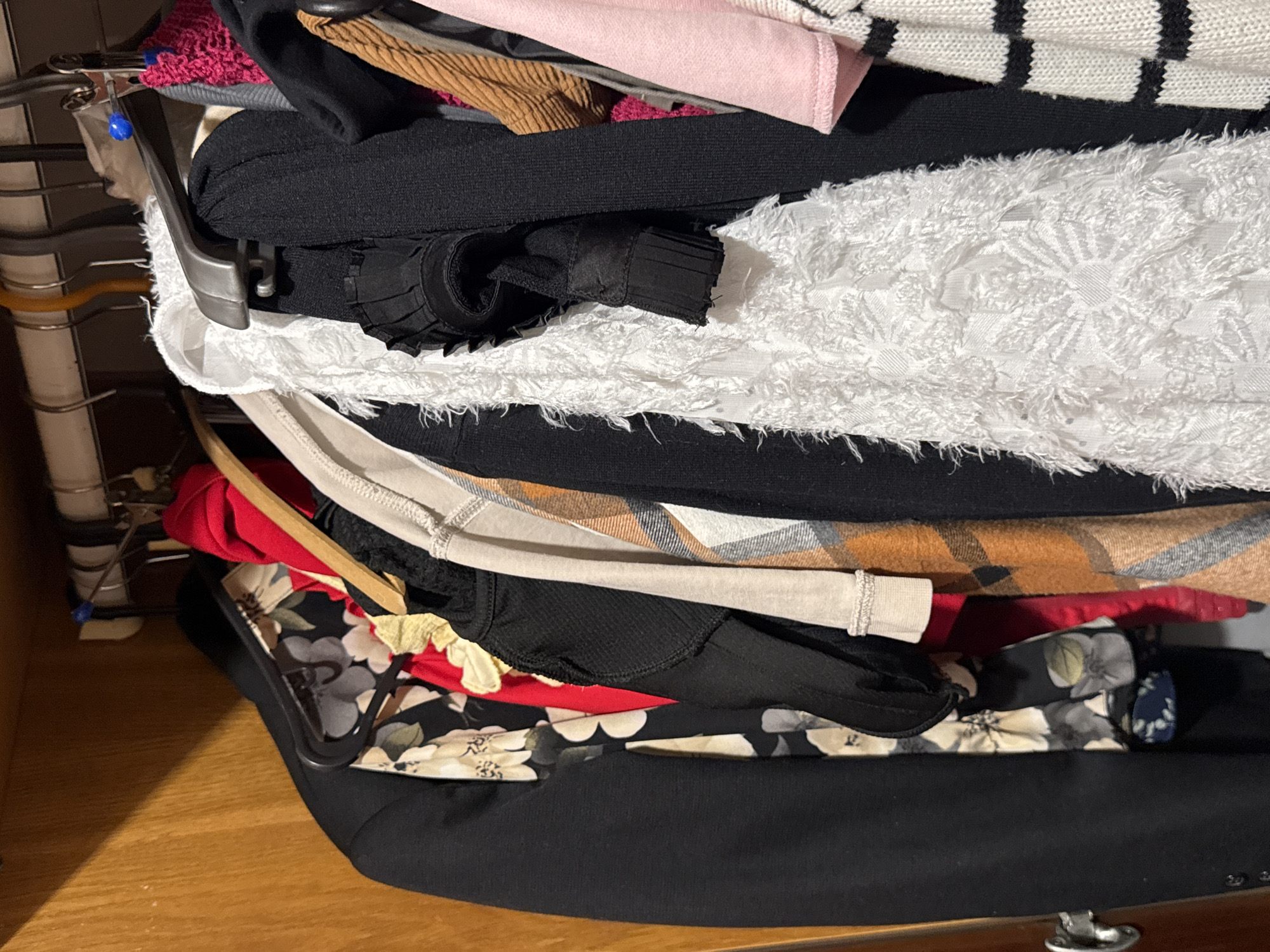 The image shows a close-up view of a closet filled with various clothing items hanging on hangers. The closet appears to be made of wood. From left to right, a black blazer is visible, followed by a floral print top. Next is a red garment, then a black top with a textured pattern, and a light-colored shirt with a plaid pattern. A white, textured garment is prominently displayed in the center, with ruffles or fringe. To its right, another black garment with textured sleeves is seen, followed by a brown ribbed garment and a pink garment. Finally, a striped sweater is partially visible on the far right. The hangers are a mix of black plastic and silver metal. There are no people or animals in the image. The lighting suggests an indoor setting. The context of Яромінскі сельскі Савет, Belarus, is not visually present in the image.