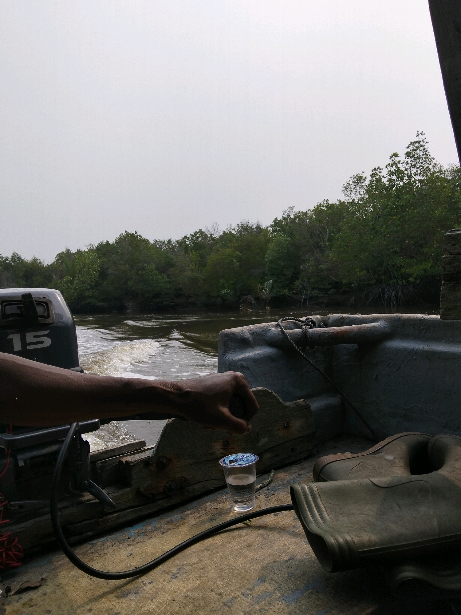 A person's arm and hand are visible in the foreground of the image, resting on the edge of a boat. The hand is clenched. To the left of the arm is a grey outboard motor with the number "15" in white. To the right of the arm is a clear plastic cup with a blue lid, containing a clear liquid. Further to the right, a pair of green rubber boots are resting on the boat's floor. The boat is on a body of water, with a wake trailing behind. Dense green foliage lines the distant shore. The sky is overcast. This scene is consistent with being in or near Kembung Luar, Indonesia, a coastal region known for its mangrove forests and waterways, often traversed by small boats.