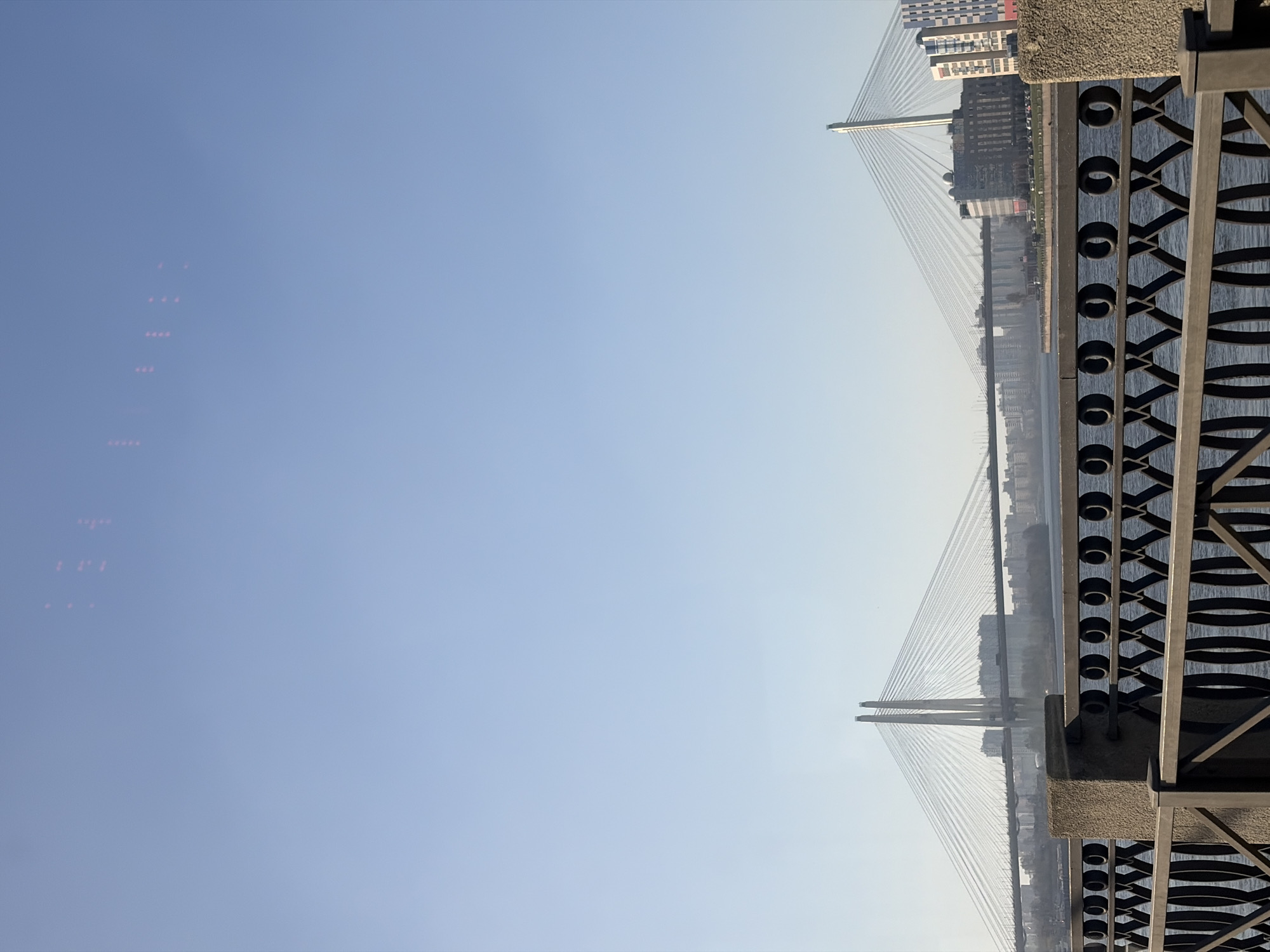 The image is an eye-level, vertical shot of the Cable-stayed Bridge (Bolshoy Obukhovsky Bridge) in Saint Petersburg, Russia, as viewed from a riverside observation point.
The bridge is the central subject, rendered in a slightly hazy atmospheric perspective. Its primary visual elements include two towers, numerous supporting cables, and the road surface. Buildings form the bridge's backdrop on either end.
The right side of the image includes a section of decorative metal railing, the repetitive circular patterns of which add texture and a foreground element. The river’s surface appears between the railing.
The vast majority of the frame is occupied by a cloudless, light blue sky, indicating daylight conditions.