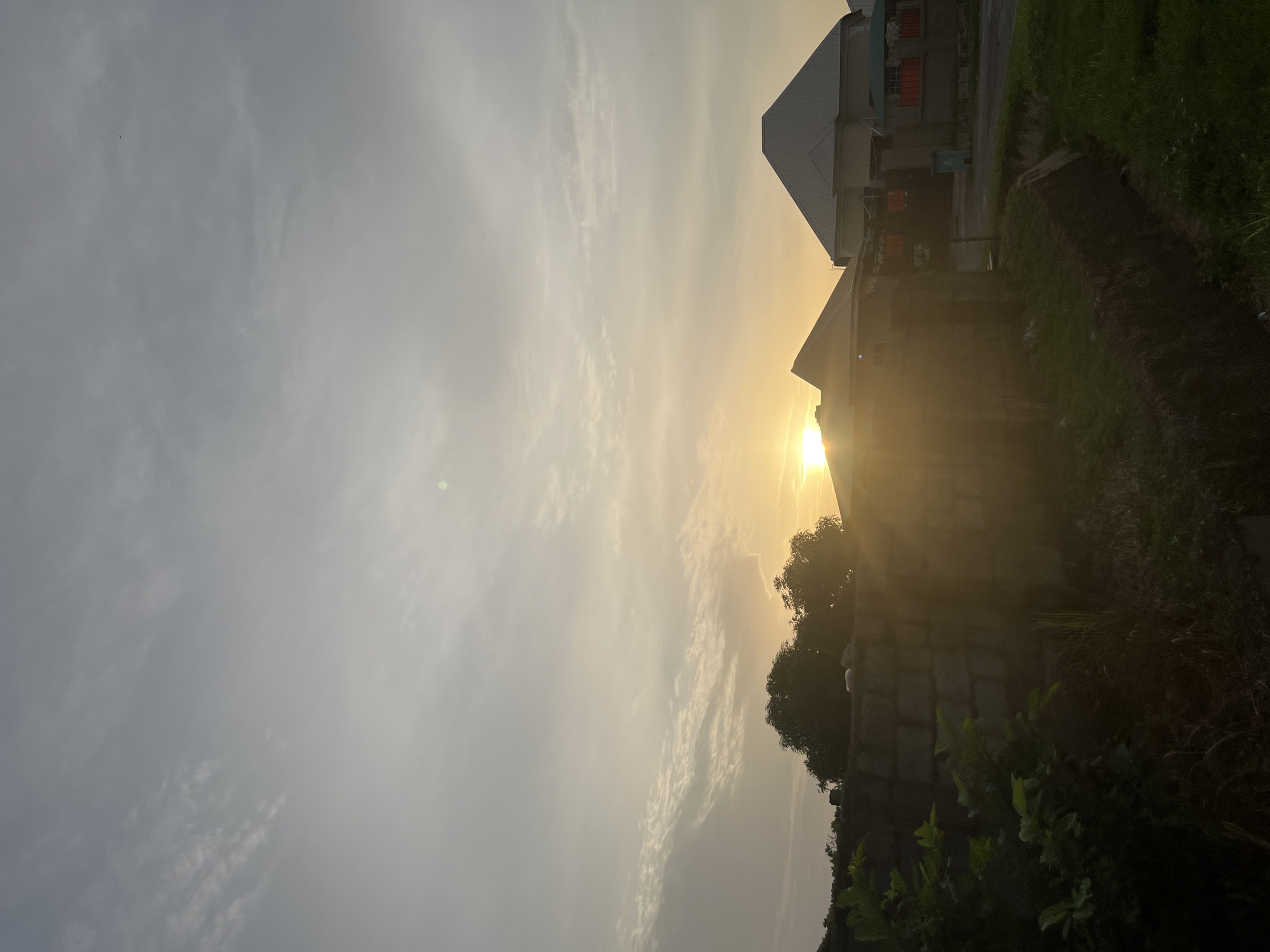 The image depicts a residential scene in Abuja, Nigeria, during either sunrise or sunset. The sky is overcast with scattered clouds, tinged with the warm hues of the sun peeking through. In the foreground, a low, rough-hewn brick wall runs horizontally across the frame, partially obscured by dense green foliage and dry grass. Behind the wall, several modern-style houses with angular roofs are visible. One house, to the right, is particularly prominent with its large, grey, pitched roof. Sunlight strongly illuminates the scene, creating lens flare and casting long shadows. The environment appears somewhat unkempt in the immediate foreground, with overgrown grass and a visible drainage channel. The overall impression is a quiet, residential area bathed in the soft, warm light of dawn or dusk.