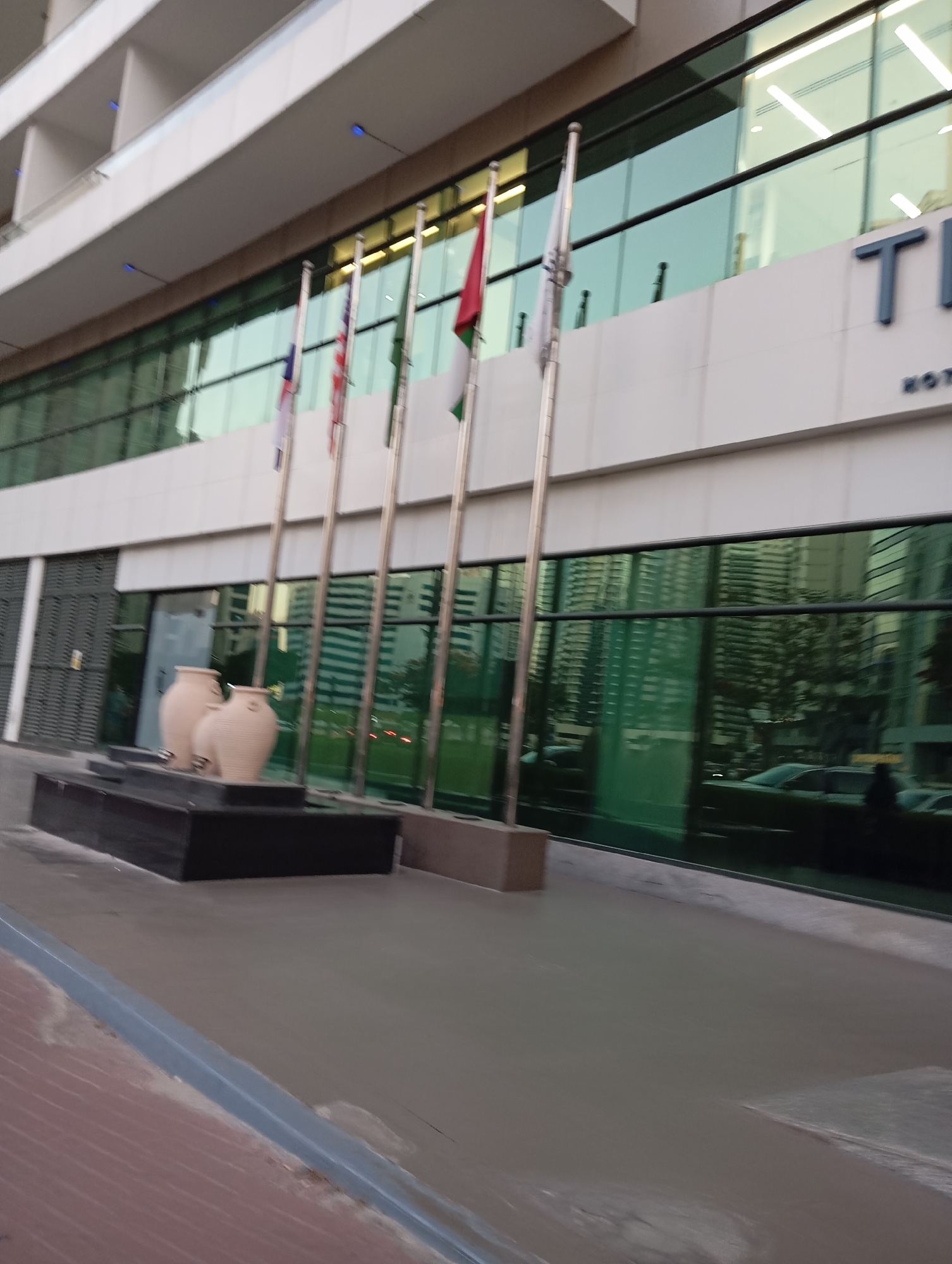 The image shows the exterior of a modern hotel building with large glass windows in Dubai, United Arab Emirates. The building has multiple levels and a white facade. In front of the building, there are several flagpoles with flags of various countries, including what appears to be the flags of the United States, the United Kingdom, and other nations. Two large, beige decorative urns are placed on a dark rectangular base in the foreground. The glass windows reflect the surrounding buildings and greenery. The ground in front of the building is paved with light-colored concrete and has a brick-like pattern in the lower left corner. The sky is not visible in the frame. The overall impression is that of a well-maintained commercial property in a contemporary urban setting.